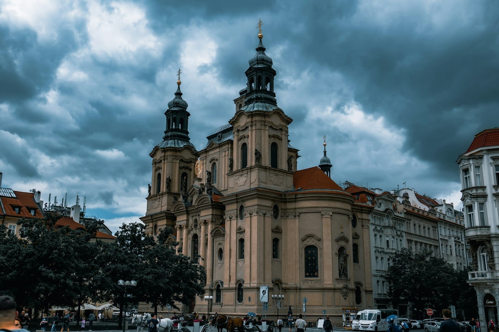 St.-Nikolaus-Kirche in der Prager Altstadt mit barocken Türmen und reich verzierter Fassade unter dramatisch bewölktem Himmel mit städtischem Treiben im Vordergrund.