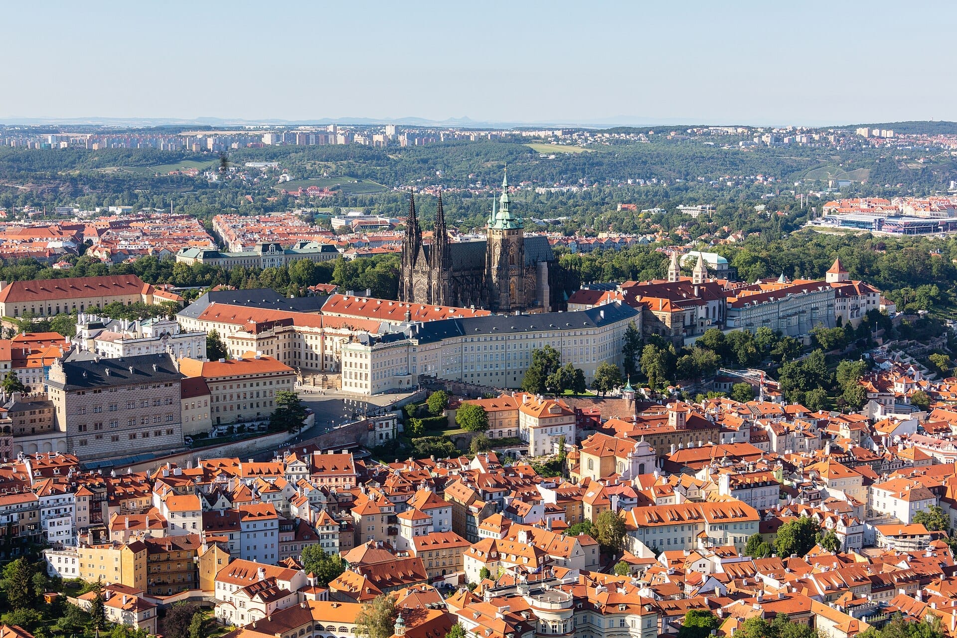 Luftaufnahme der Prager Burg mit ihrer gotischen Kathedrale, umgeben von Gebäuden mit roten Dächern und grünen Hügeln unter einem klaren blauen Himmel.