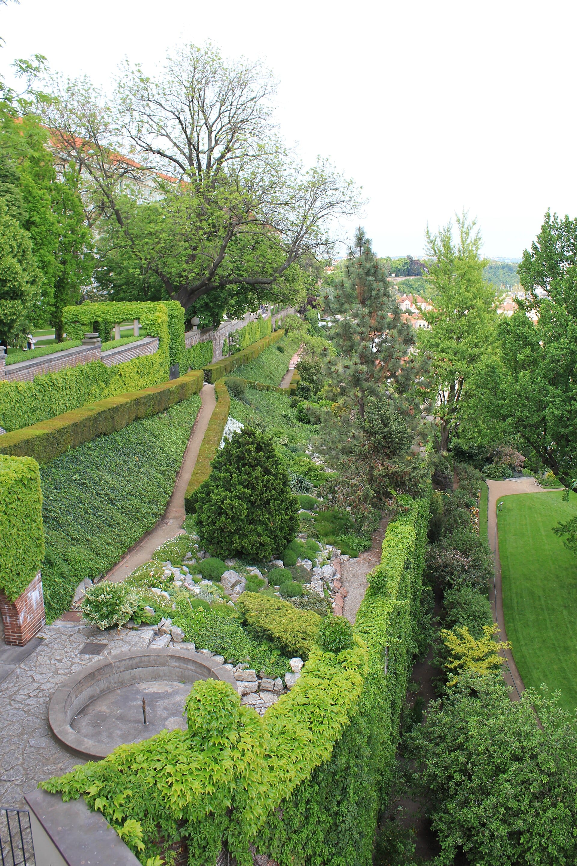 Blick auf die Südgärten der Prager Burg mit terrassenförmigem Mauerwerk, üppigen grünen Hecken, verschlungenen Wegen und Zierpflanzen unter einem hellen Himmel.