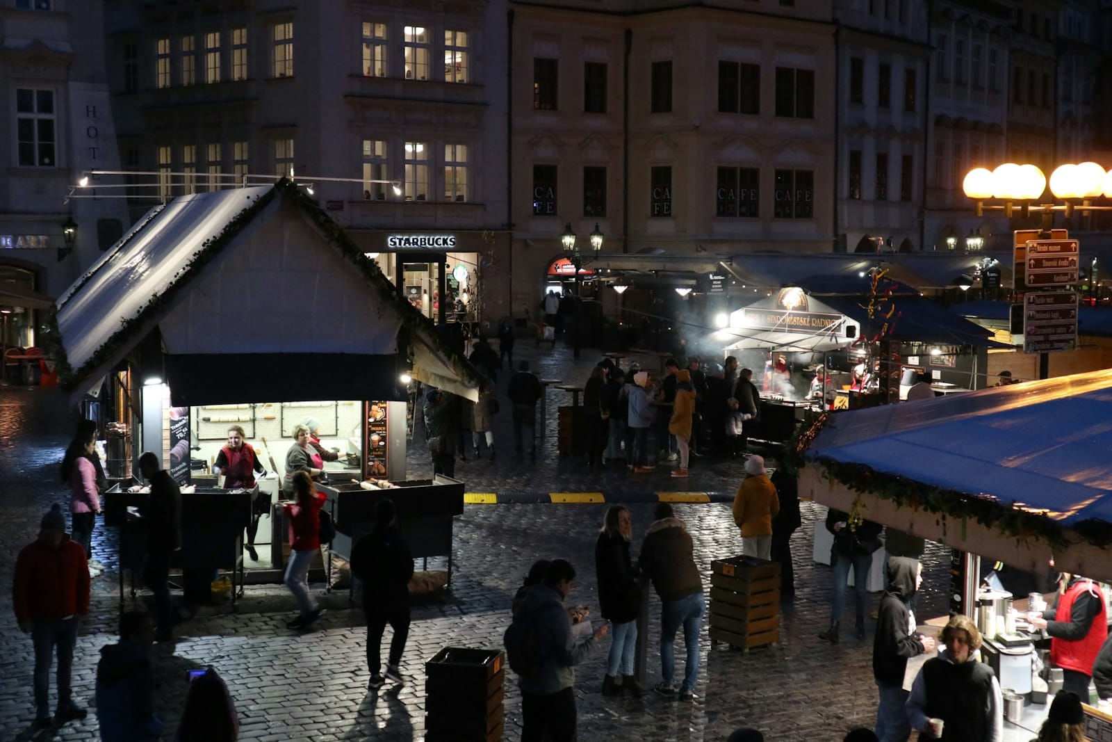 Weihnachtsmarktstände in Prag bei Nacht, mit Menschen, die Essen und Getränke kaufen, Kopfsteinpflasterstraßen und festlichen Lichtern.