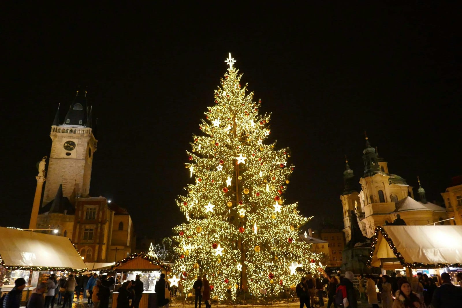 Großer, mit Lichtern geschmückter Weihnachtsbaum auf dem Altstädter Ring in Prag, mit festlichen Marktständen und historischen Türmen bei Nacht.