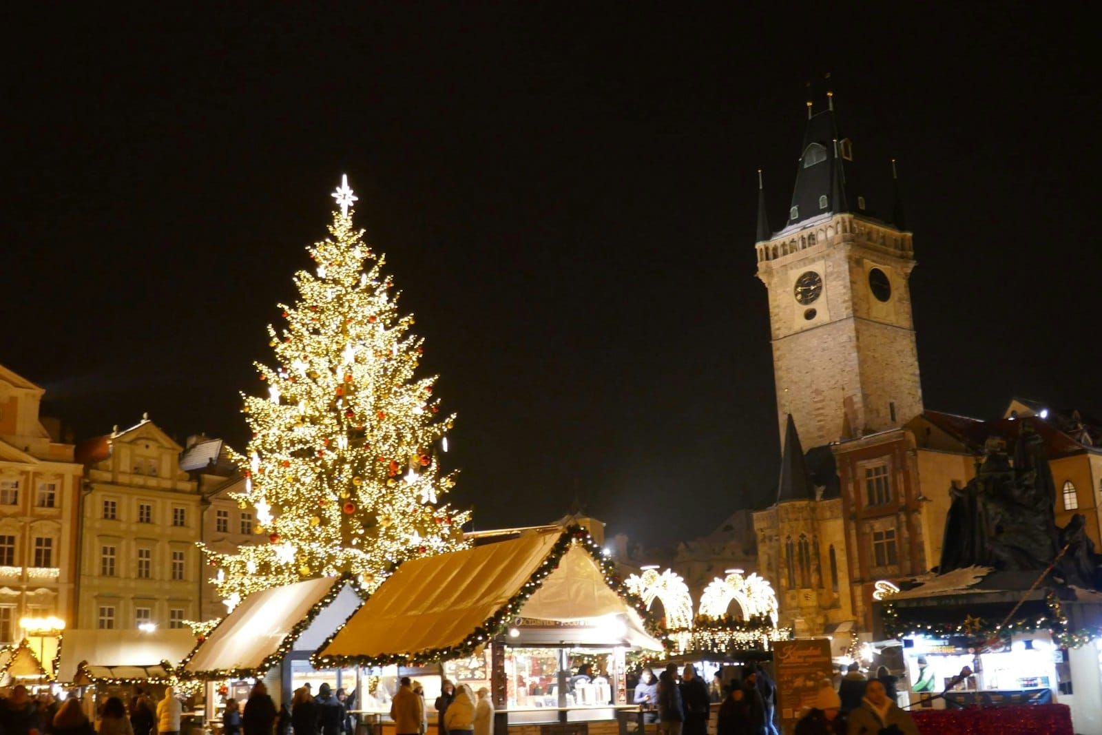 Nächtliche Szene des Weihnachtsmarkts auf dem Altstädter Ring in Prag mit einem großen geschmückten Baum, festlichen Ständen, Menschenmassen und dem sichtbaren Altstädter Rathausturm.