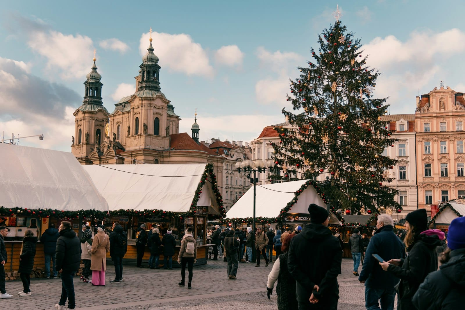 Weihnachtsmarkt auf dem Altstädter Ring in Prag mit festlichen Buden, einem großen geschmückten Weihnachtsbaum, schlendernden Besuchern und historischen Kirchtürmen vor blauem Himmel.