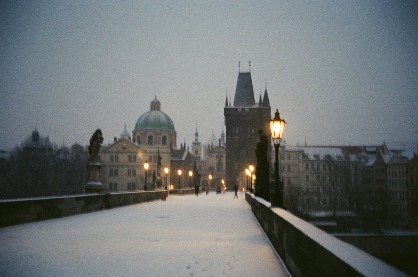 Schneebedeckte Charles Bridge in Prag in der Abenddämmerung mit historischen Gebäuden und leuchtenden Straßenlaternen im Winter.