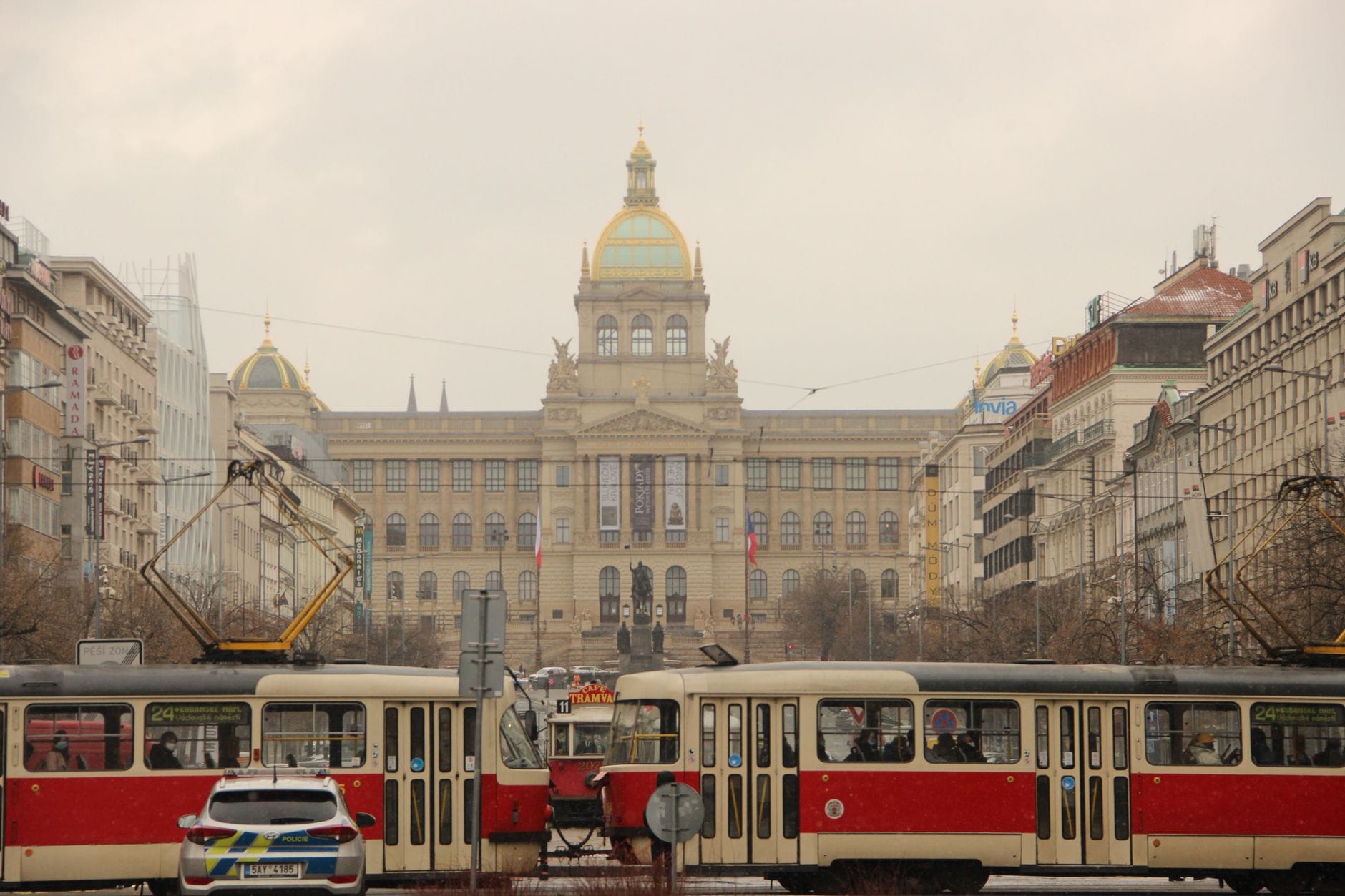 Klassische Prager Straßenbahnen und ein Polizeiauto, die vor dem ikonischen Nationalmuseum am Wenzelsplatz mit historischer Stadtarchitektur vorbeifahren.