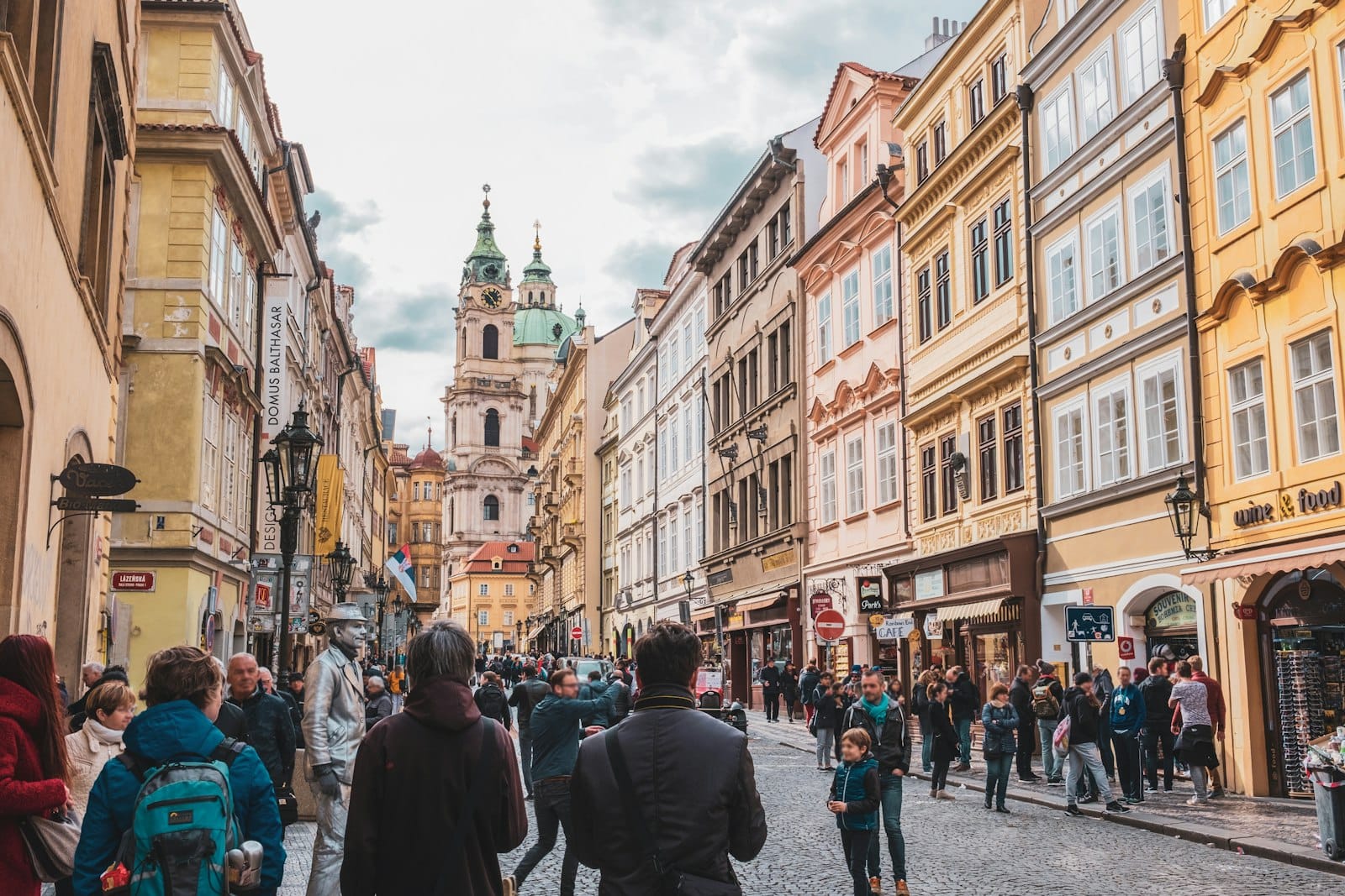 Menschenmassen spazieren entlang einer berühmten Kopfsteinpflasterstraße im historischen Zentrum von Prag, gesäumt von pastellfarbenen Gebäuden und Cafés, mit einer Kirche in der Ferne.