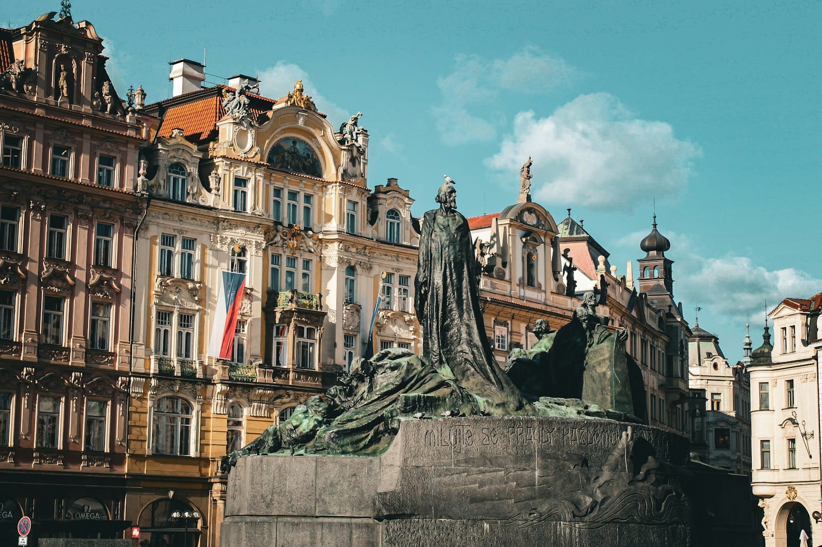 Große Bronzestatue auf einem lebhaften Prager Platz mit prachtvollen Pastellgebäuden und strahlend blauem Himmel im Hintergrund.