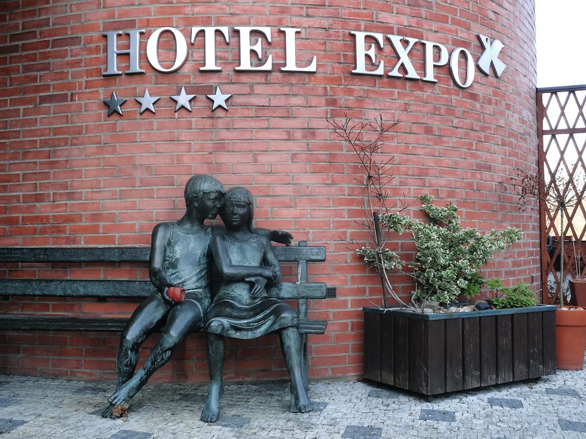 Bronzestatue eines Paares, das auf einer Bank vor dem Hotel Expo in Prag sitzt, vor einer geschwungenen roten Backsteinmauer und Topfpflanzen.