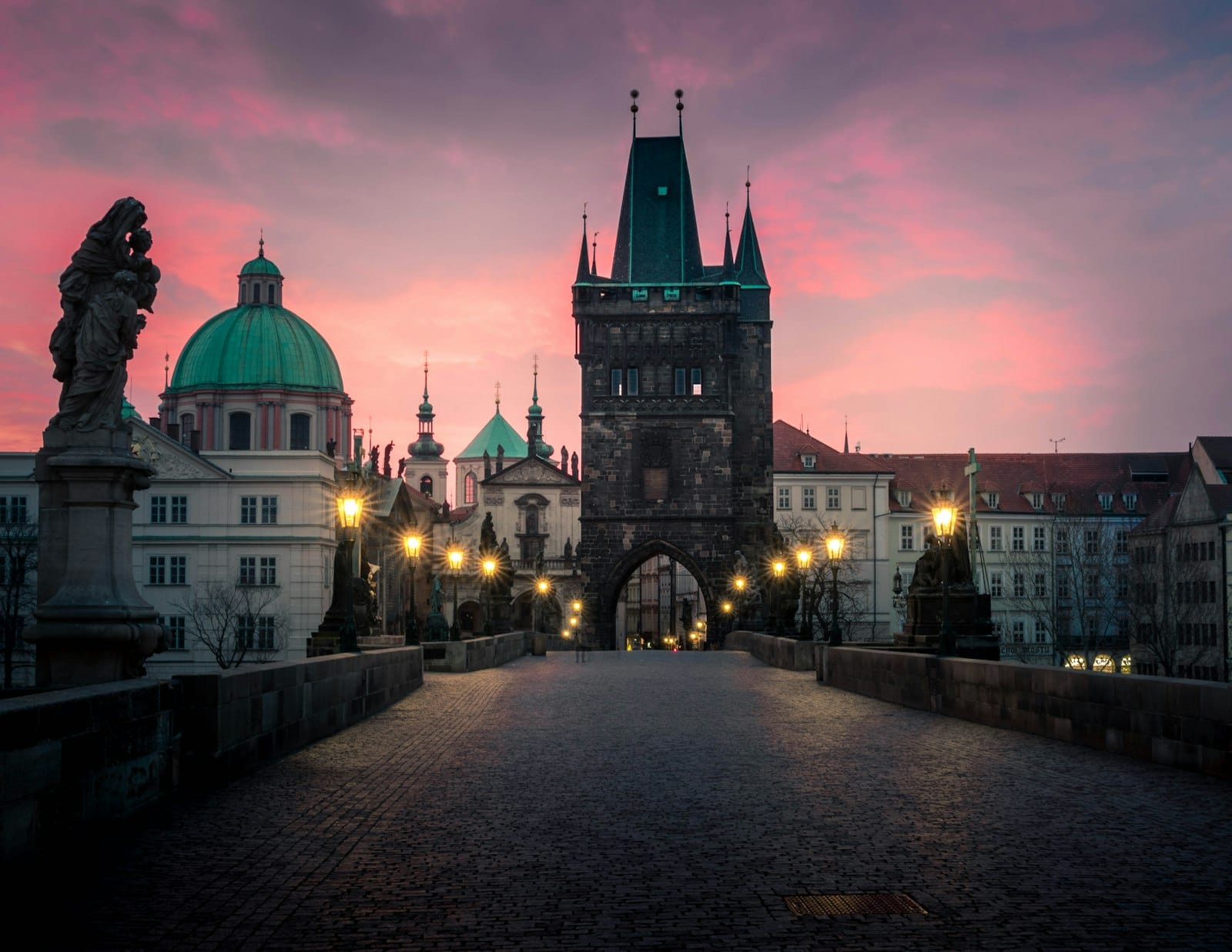 Die Karlsbrücke in Prag bei Sonnenaufgang, leerer Kopfsteinpflasterweg, historische Statuen und Altstadttürme vor einem rosa Himmel.