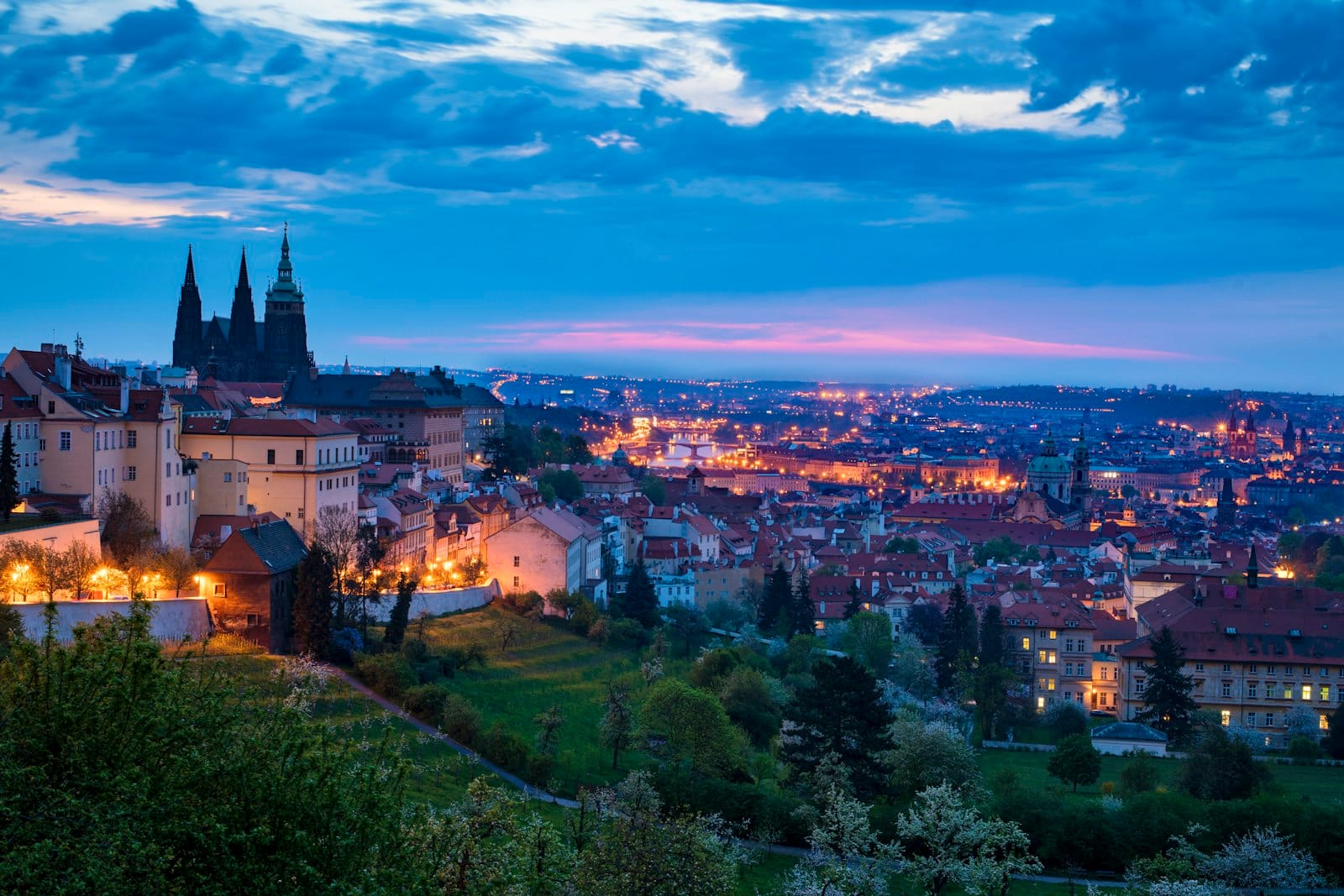 Abendliches Panorama über Prag mit leuchtenden Stadtlichtern, grünem Hang und historischen Gebäuden unter einem dramatisch blau-lila Himmel.