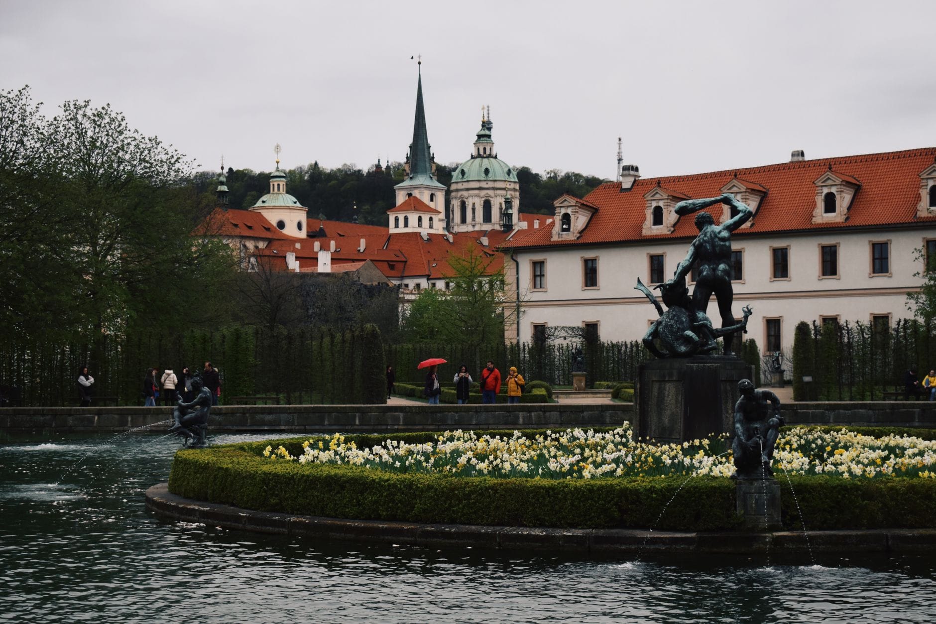 Brunnen mit Statuen, umgeben von blühenden Gärten und historischer Prager Architektur mit Kirchtürmen im Hintergrund.