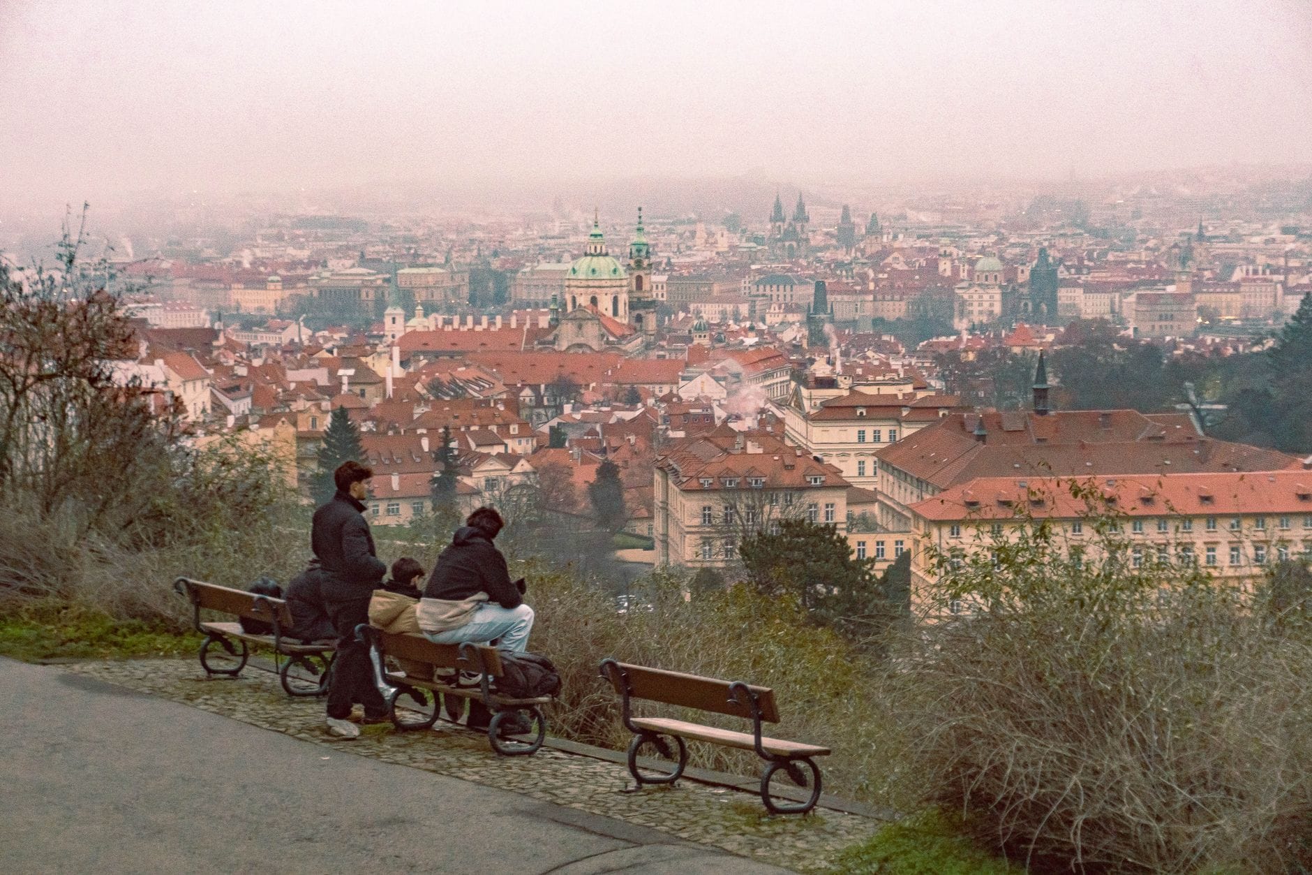 Zwei Personen sitzen auf Bänken an einem Aussichtspunkt auf einem Hügel mit Panoramablick auf Prags Altstadt und entfernte Sehenswürdigkeiten.