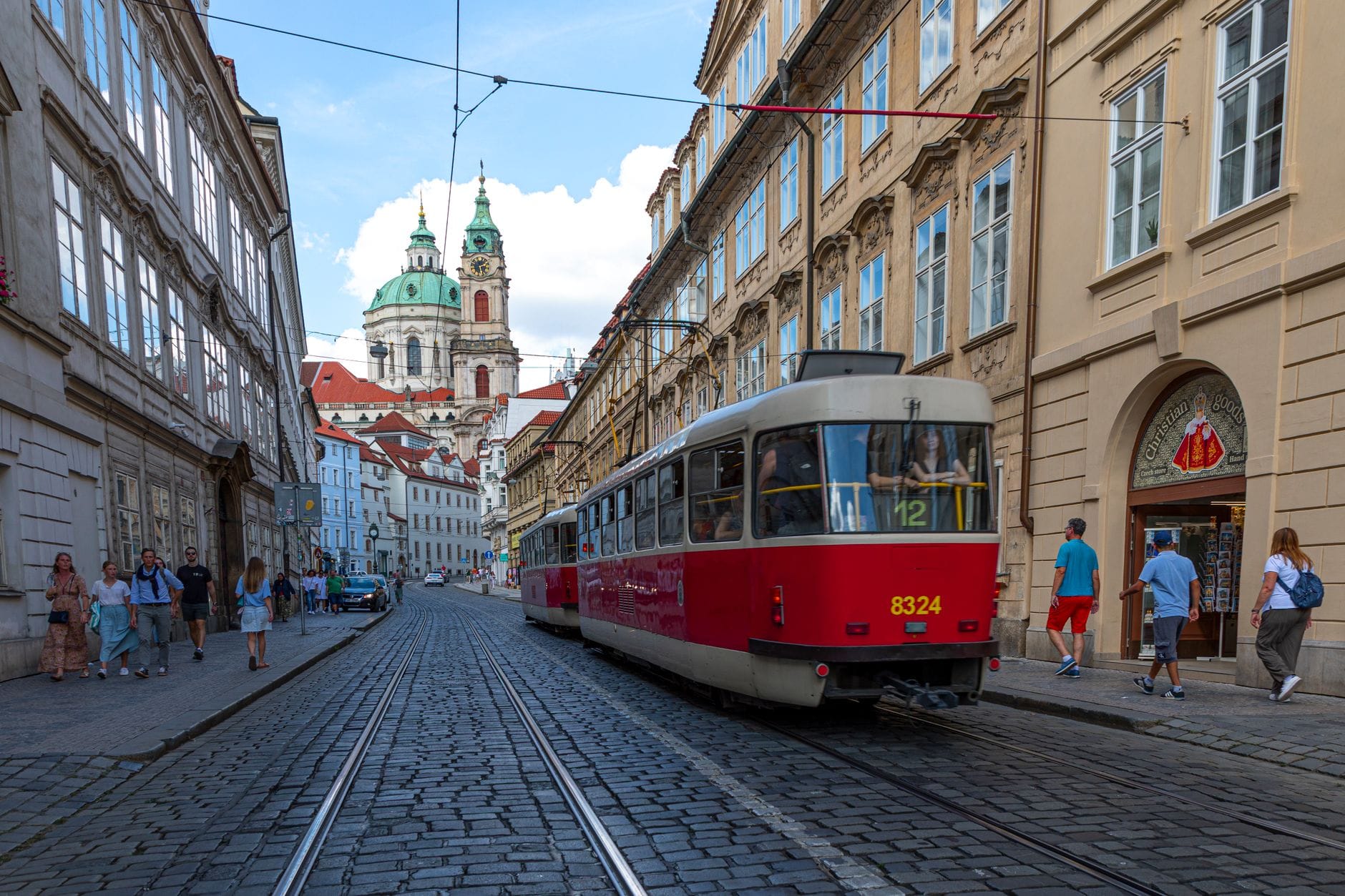 Strada del centro di Praga con tram storico, persone che camminano e la Chiesa di San Nicola sullo sfondo in una giornata limpida e soleggiata.