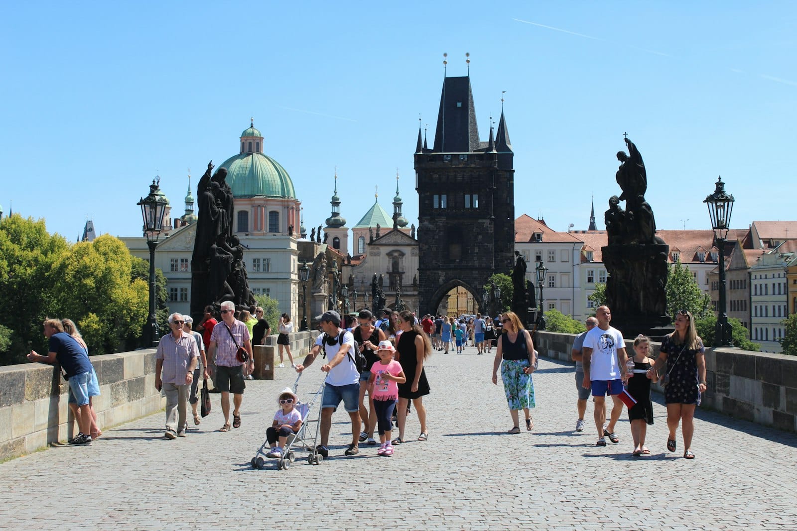 Folle di persone in abiti estivi camminano sul Ponte Carlo a Praga sotto un cielo azzurro limpido e un sole splendente.