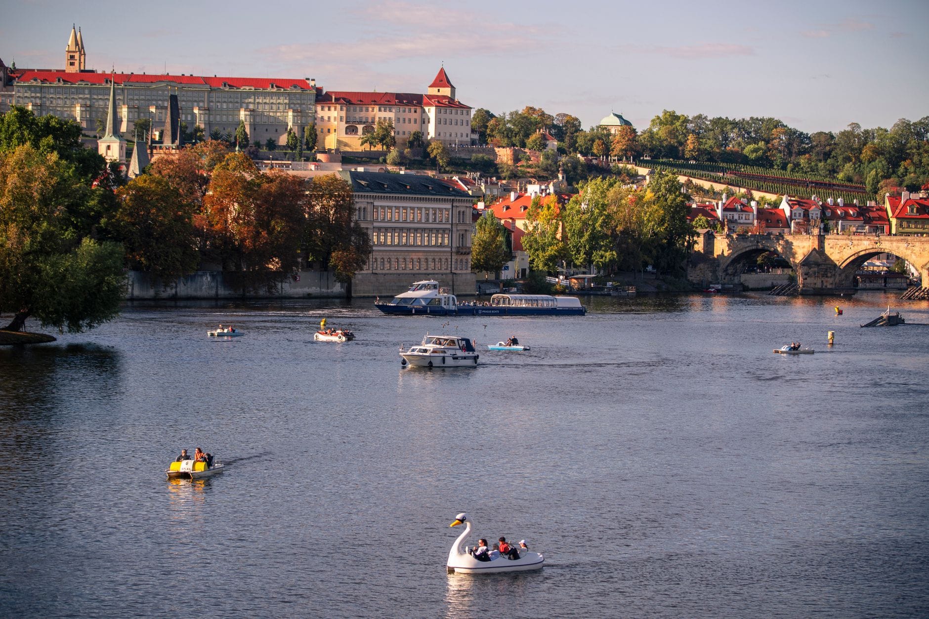 Vista del Castello di Praga e del Ponte Carlo con barche e pedalò sul fiume Vltava in una giornata di sole.