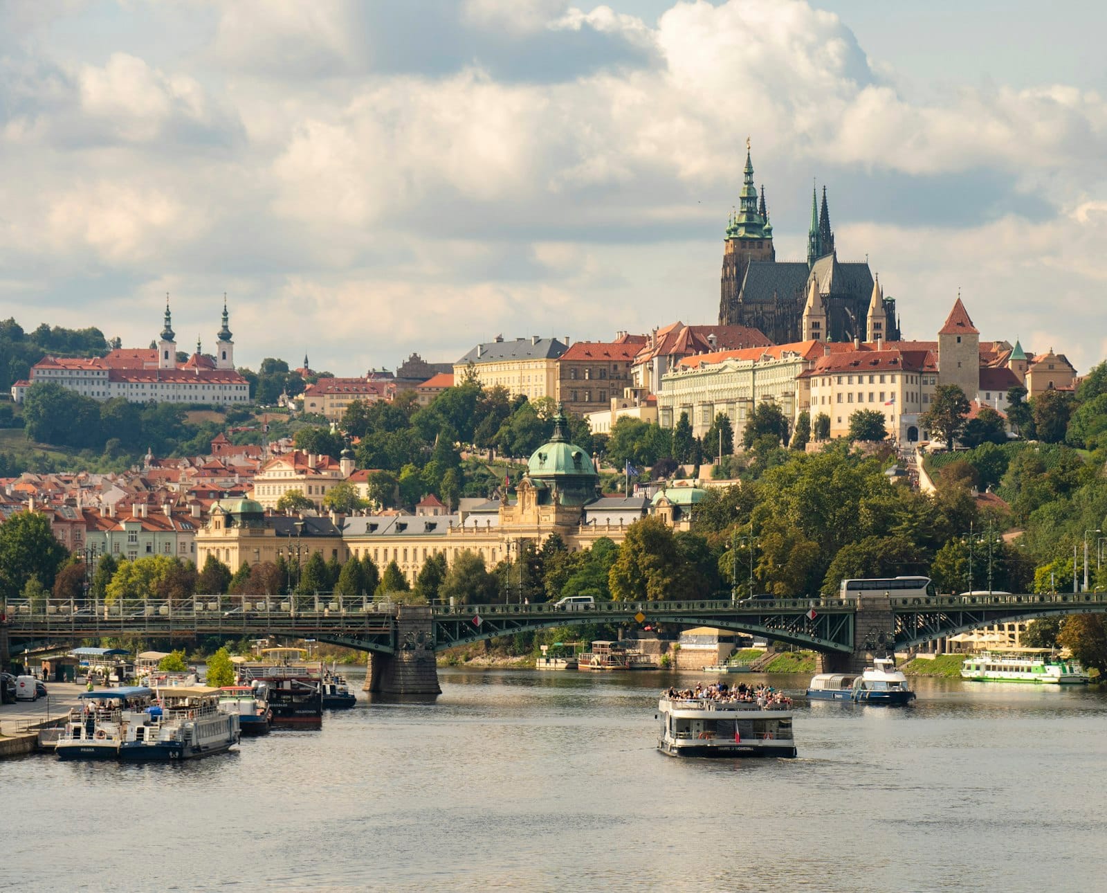 Vista del Castello di Praga, della Cattedrale di San Vito, del fiume e di imbarcazioni sotto un cielo estivo con rigogliosi alberi verdi in primo piano.