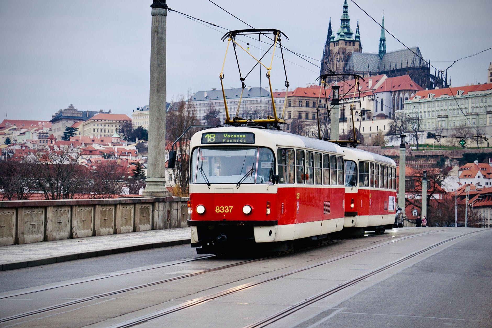 Eine klassische rot-weiße Prager Straßenbahn fährt über eine Brücke, im Hintergrund thront die Prager Burg auf einem Hügel an einem grauen Wintertag.