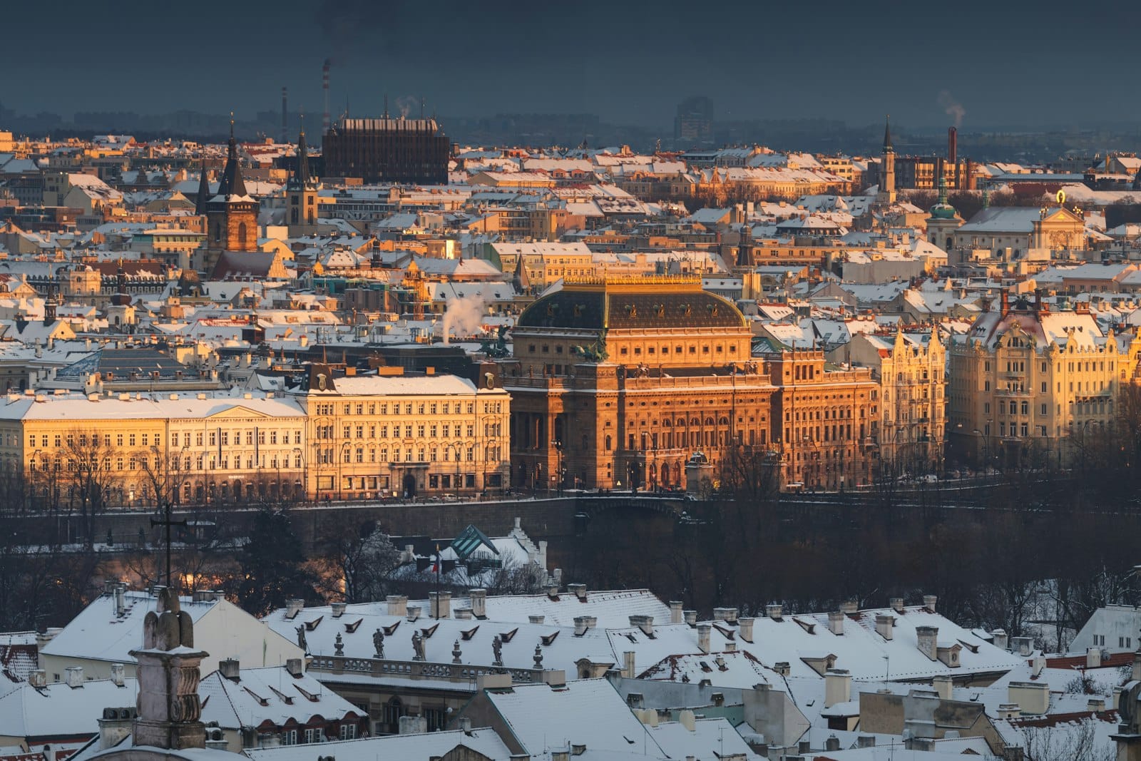 Winterpanorama von Prag mit schneebedeckten Dächern und Straßen, goldenes Sonnenlicht beleuchtet das historische Stadtzentrum vor einem stimmungsvoll grauen Himmel.