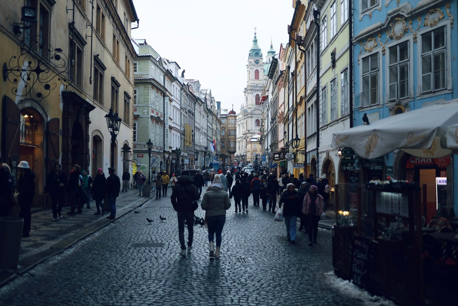 Menschen in Winterkleidung gehen auf einer Kopfsteinpflasterstraße im historischen Zentrum von Prag, umgeben von bunten alten Gebäuden.