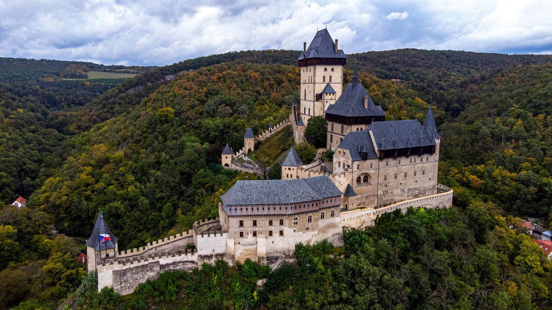 Luftaufnahme einer großen mittelalterlichen Burg, umgeben von grünen bewaldeten Hügeln unter einem bewölkten Himmel