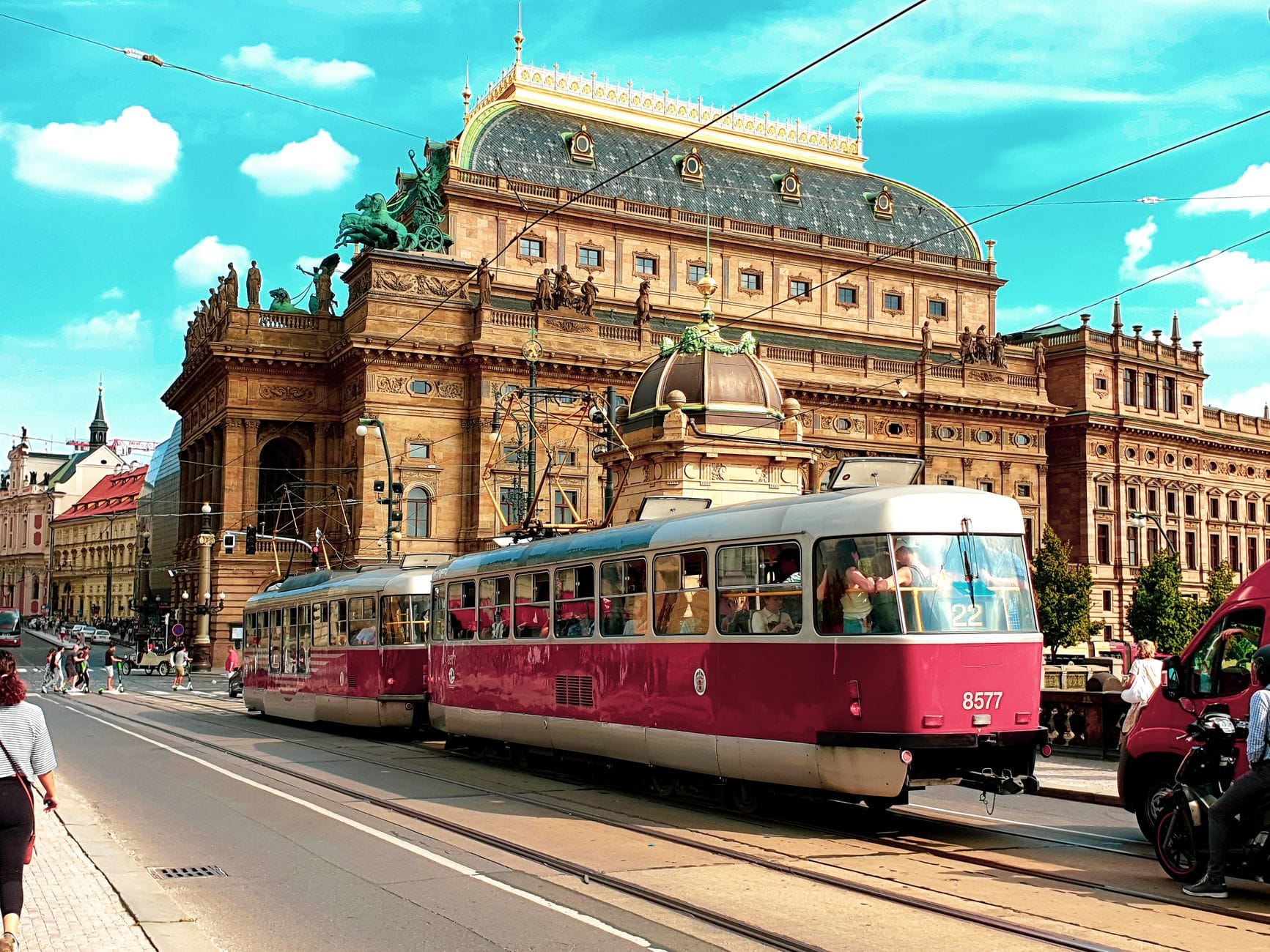 Historisches goldenes Opernhaus in Prag mit kunstvollen Dachskulpturen und roten Straßenbahnen, die auf einer sonnenbeschienenen Stadtstraße vorbeifahren.