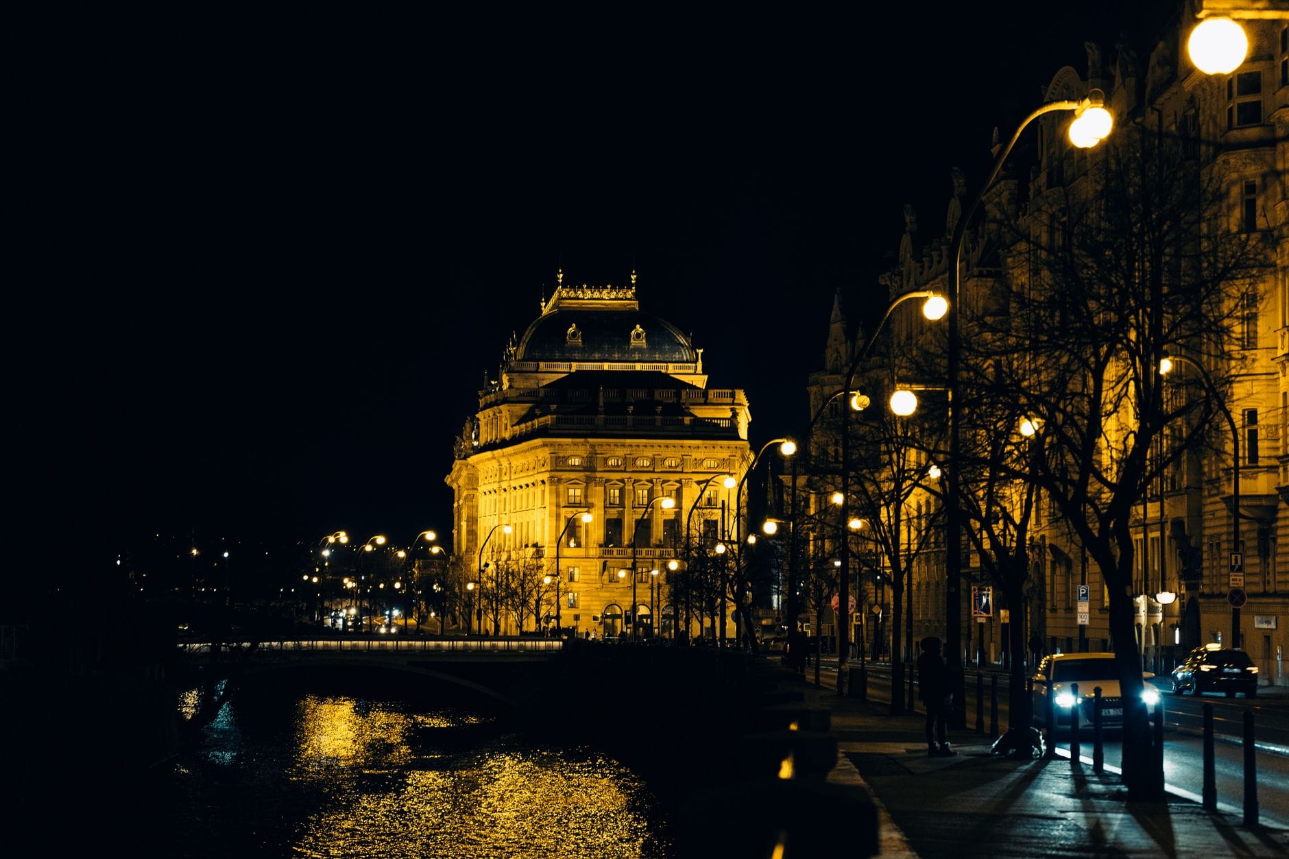 Tanawin sa gabi ng naiilawang gusali ng National Theatre sa Prague, na may mga ilaw ng kalye na nag-aanyong salamin sa Ilog Vltava at mga taong naglalakad sa paligid.