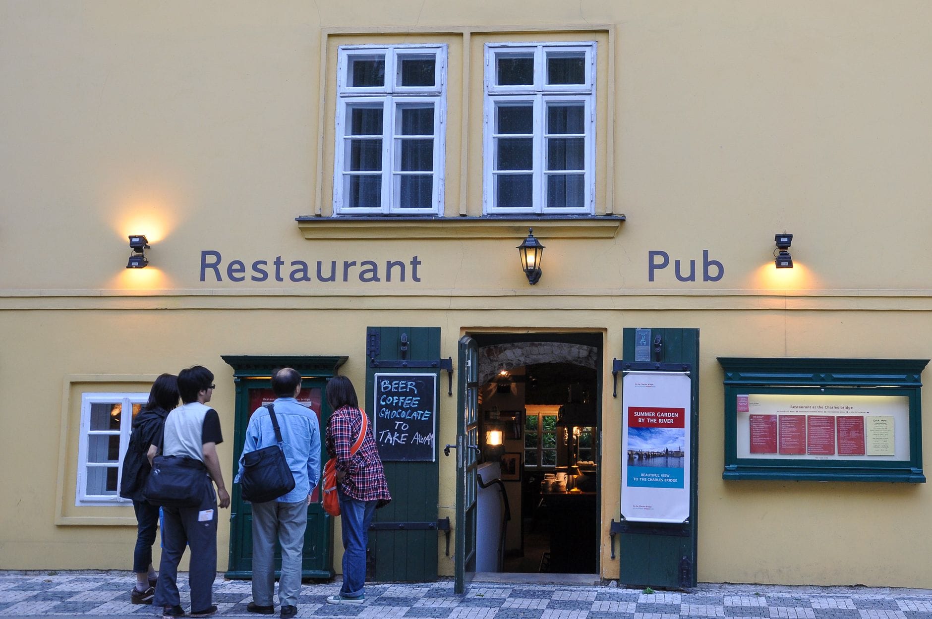 Mga taong nakatayo sa labas ng isang kaswal na restaurant at pub sa Prague, nagbabasa ng menu sa tabi ng pasukan sa isang cobblestone na bangketa.