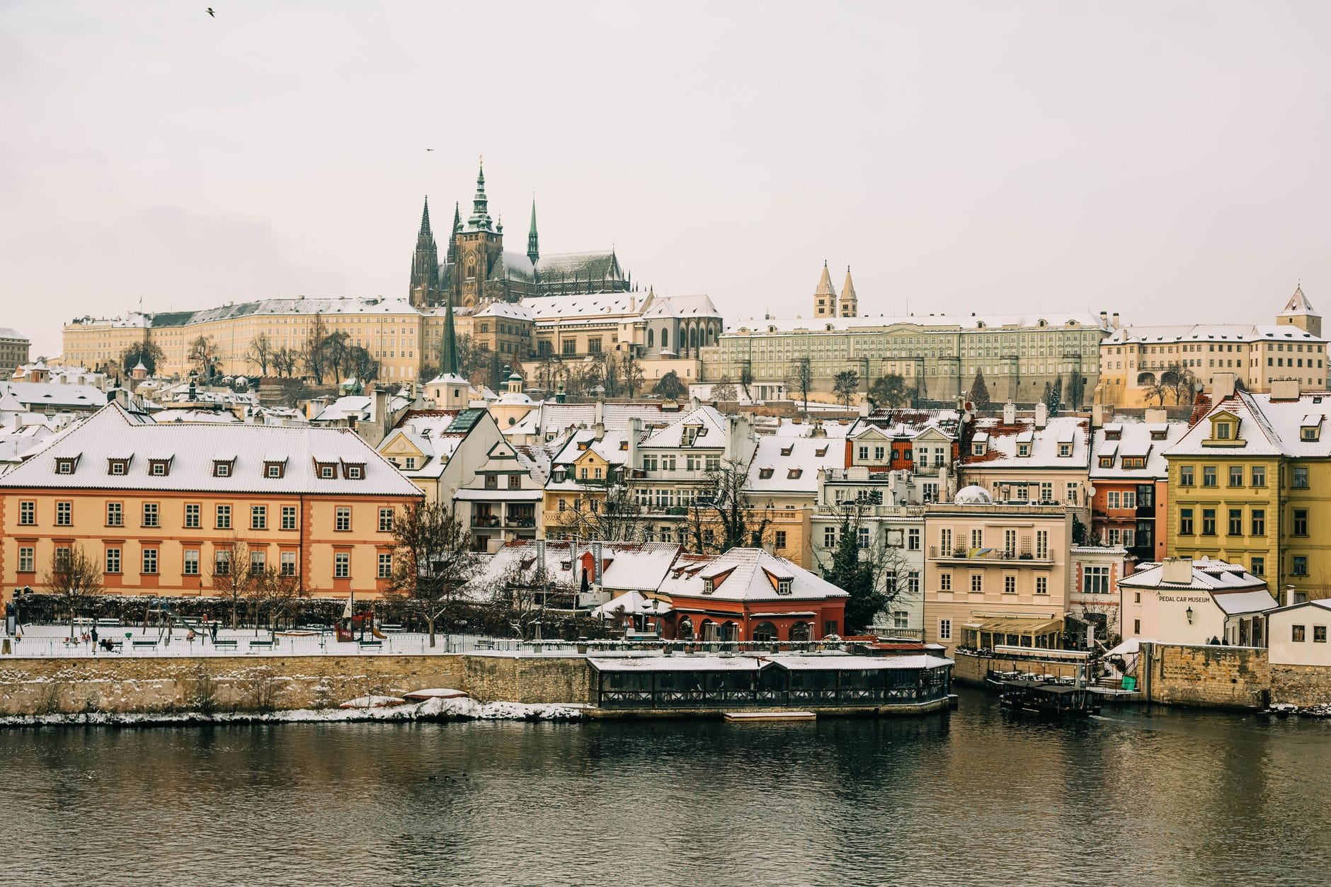 Mga bubong na niyebe at mga gusali sa tabi ng ilog sa Praha na may Praha Castle at St. Vitus Cathedral sa likod sa isang malamig na araw ng taglamig.
