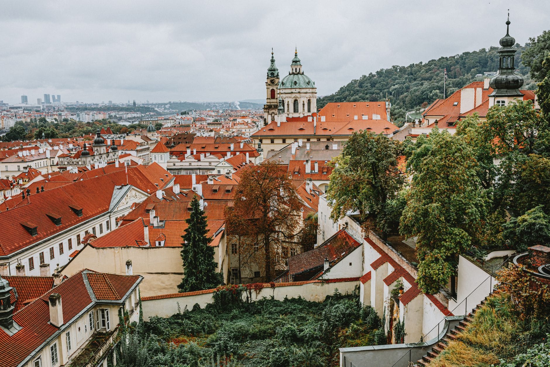 Malawak na tingin sa Malá Strana na kapitbahayan ng Prague na may mga iconic na pulang bubong, mga Baroque na gumbong simbahan, at mga berdeng burol sa likod sa ilalim ng maulap na langit.
