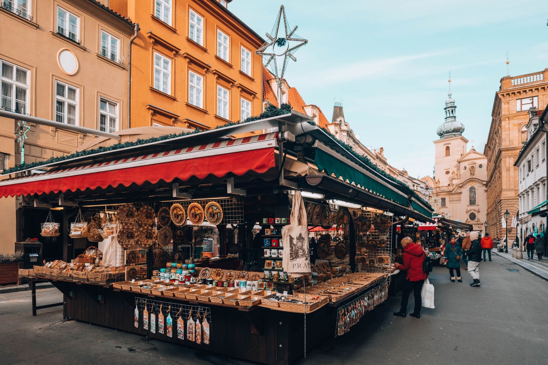 Ein belebter Outdoor-Marktstand in Prag, der lokales Kunsthandwerk und Lebensmittel verkauft, mit historischen Gebäuden und einer Kirche im Hintergrund.