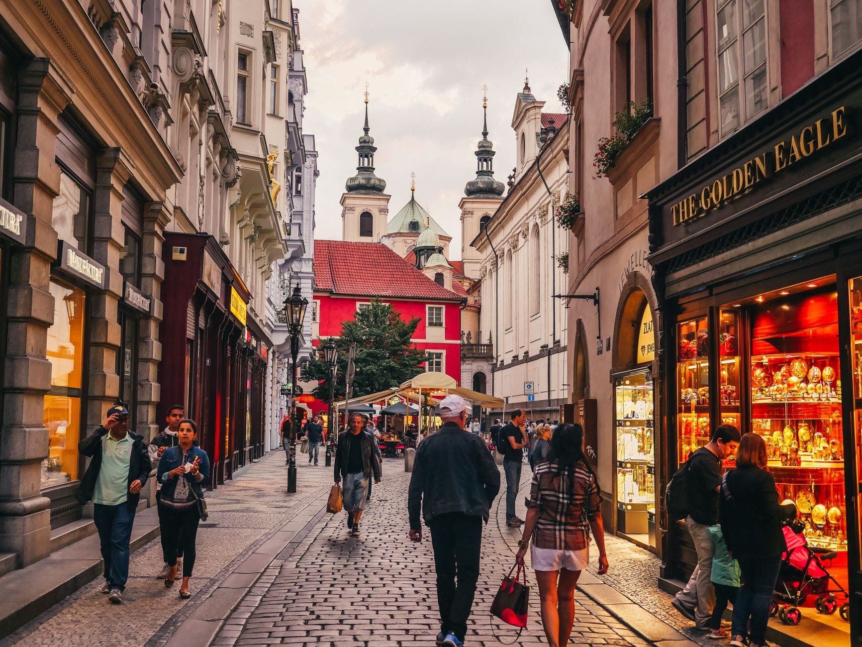 Kopfsteinpflaster-Einkaufsstraße in Prag mit eleganten Geschäften, flanierenden Menschen, historischen Gebäuden und Kirchtürmen im Hintergrund bei Sonnenuntergang.