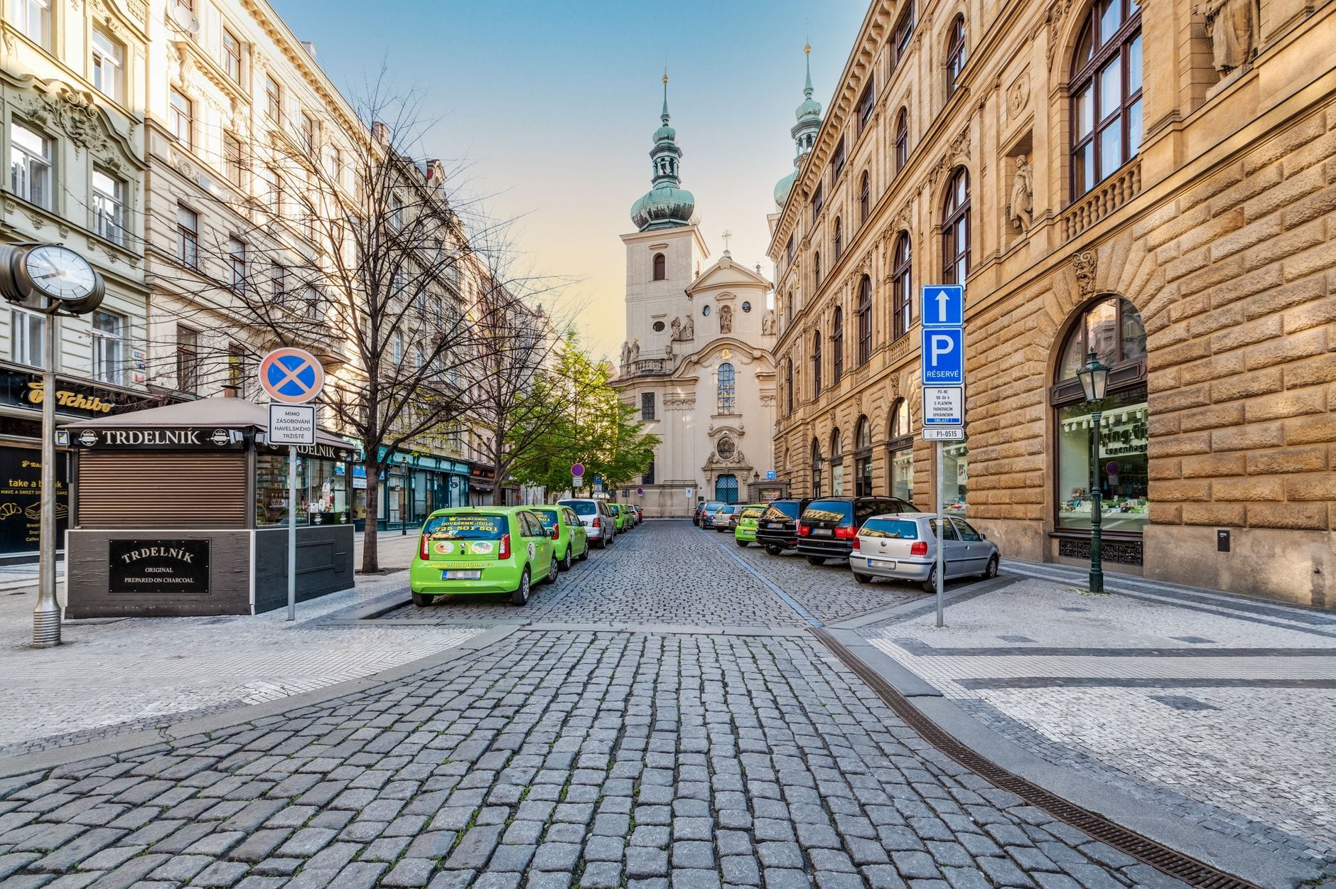 Strada acciottolata a Praga fiancheggiata da auto parcheggiate e taxi verdi, con edifici barocchi e una chiesa sullo sfondo.