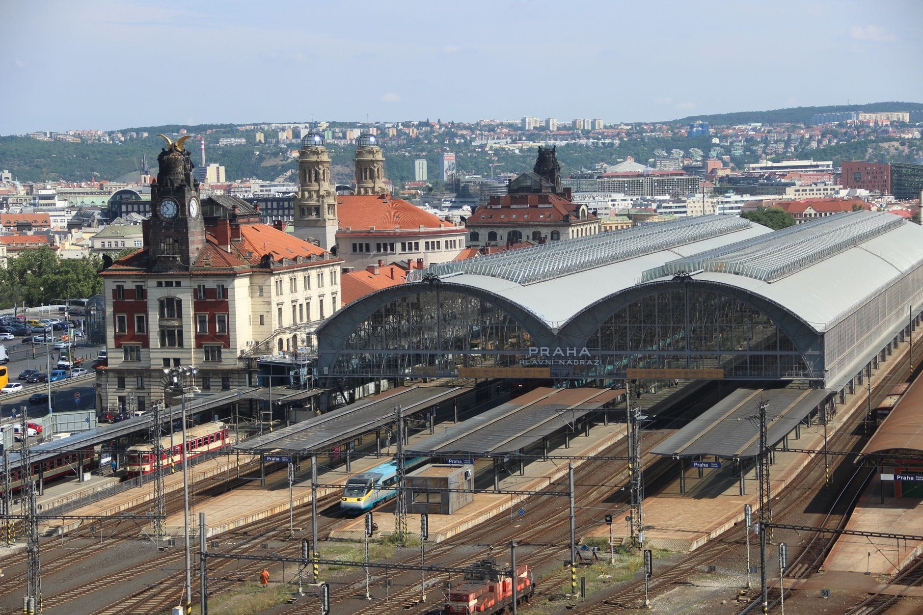 Widok z lotu ptaka na główny dworzec kolejowy w Pradze z wieloma torami, peronami, zabytkową architekturą i panoramą miasta w tle pod bezchmurnym niebem.