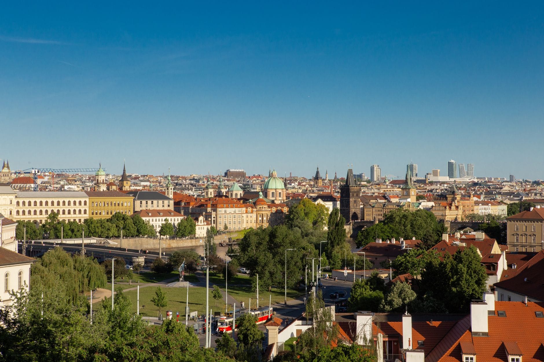 Panoramiczny widok na panoramę Pragi z czerwonymi dachami, historycznymi wieżami, zielonymi przestrzeniami i nowoczesnymi budynkami pod bezchmurnym błękitnym niebem.