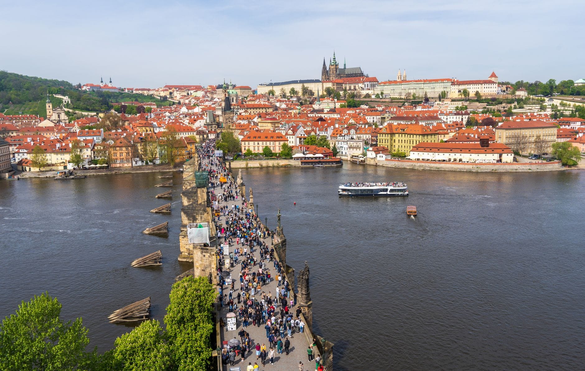 Luftaufnahme der von Menschen gefüllten Karlsbrücke, der Moldau und der Prager Burg im Hintergrund an einem sonnigen Tag.