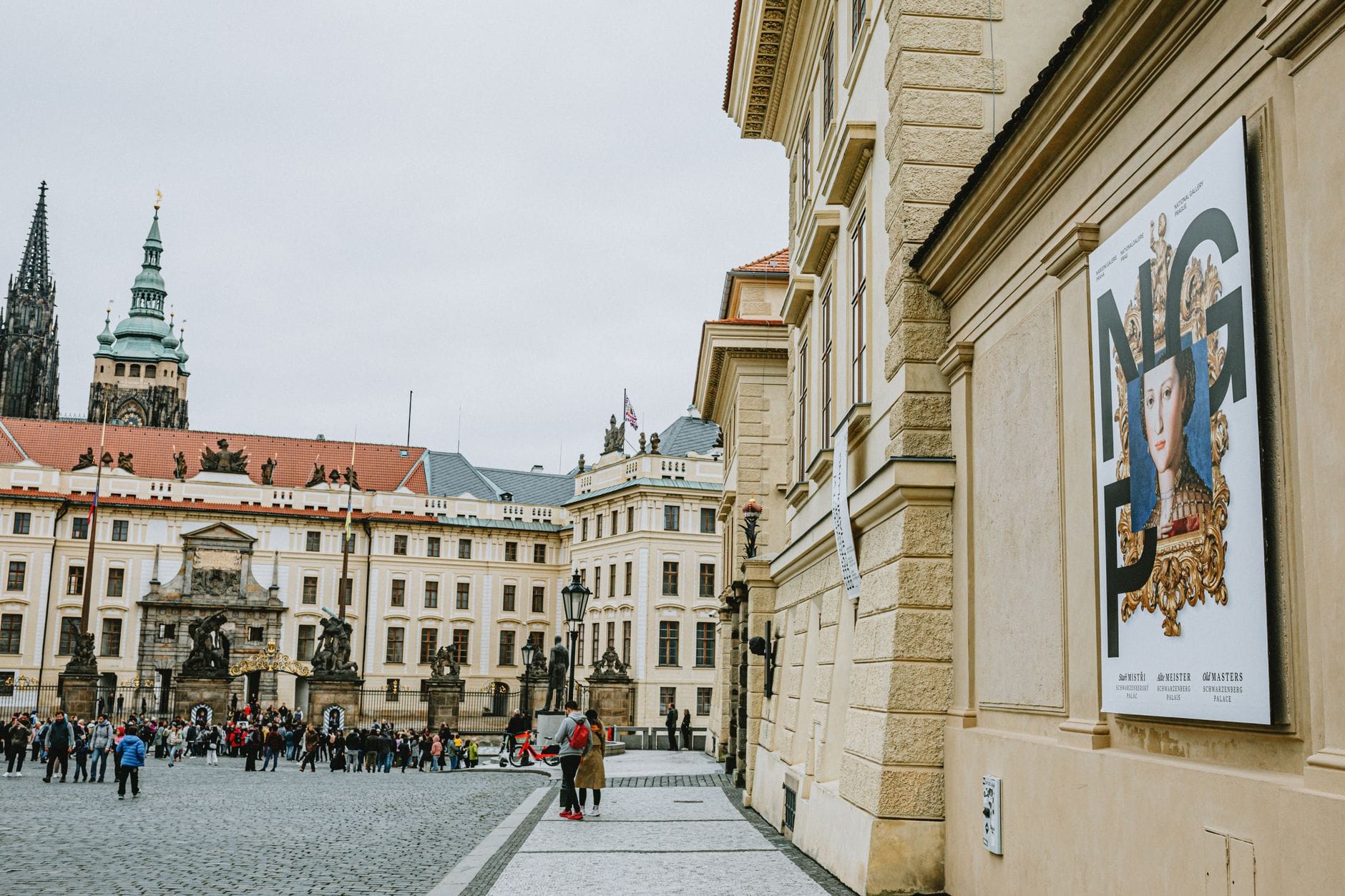 Innenhof des Burgkomplexes von Prag, mit Gruppen von Touristen, historischen Gebäuden und den im Hintergrund sichtbaren Türmen des Veitsdoms.