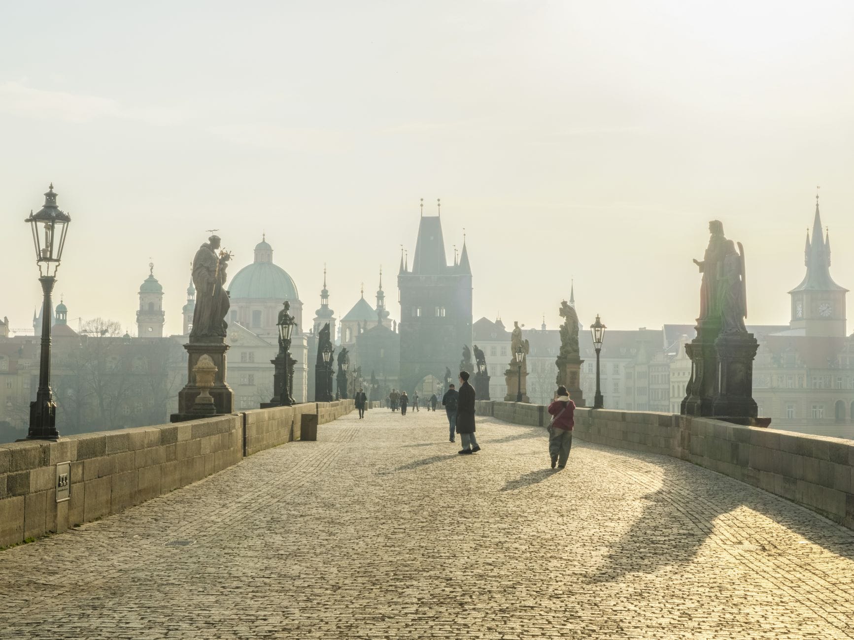 Frühmorgendliche Ansicht der Karlsbrücke in Prag mit nur wenigen Spaziergängern und sanftem Sonnenlicht, zeigt geringe Besucherzahlen.