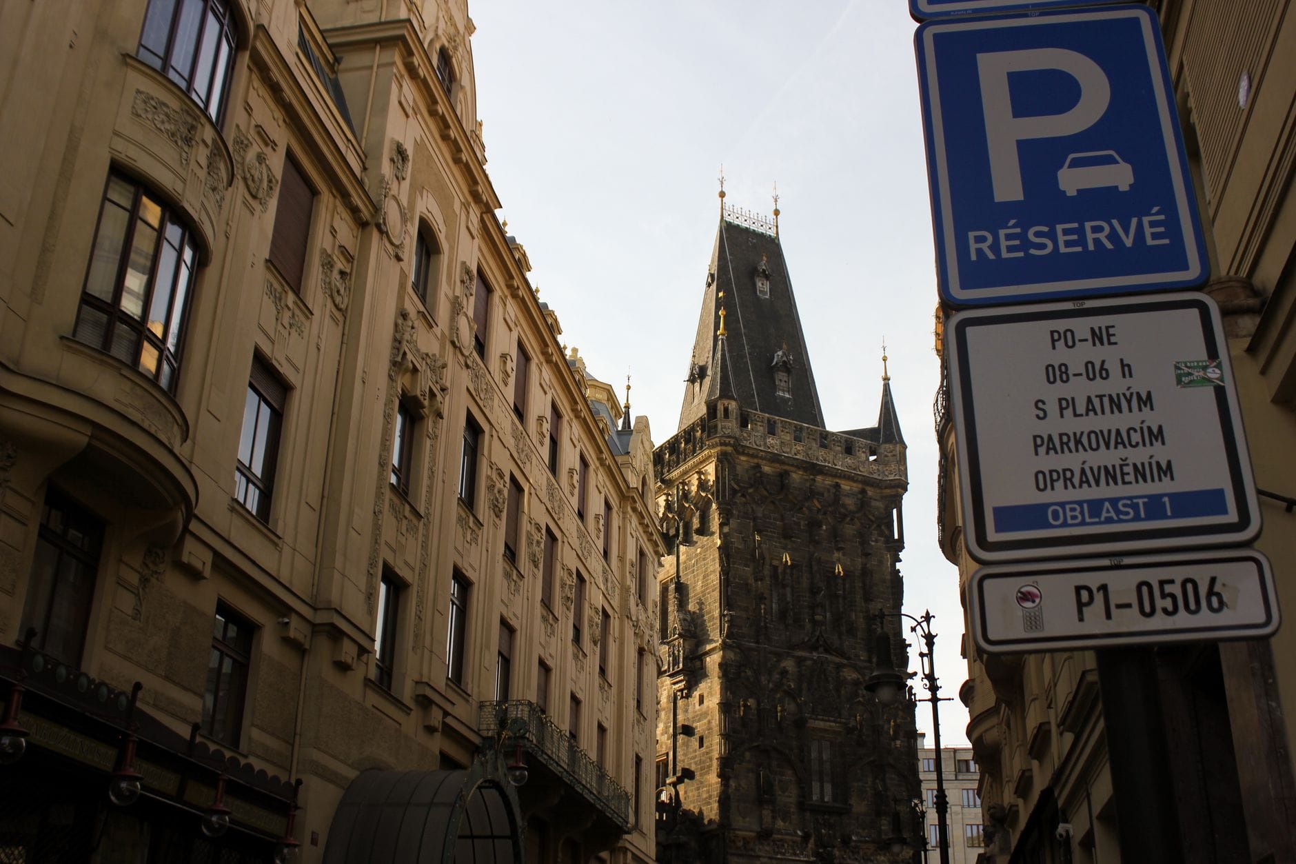 Straßenansicht mit Blick auf den gotischen Pulverturm (Prašná brána) in Prag, eingerahmt von historischen Gebäuden und einem sichtbaren Parkplatzschild.