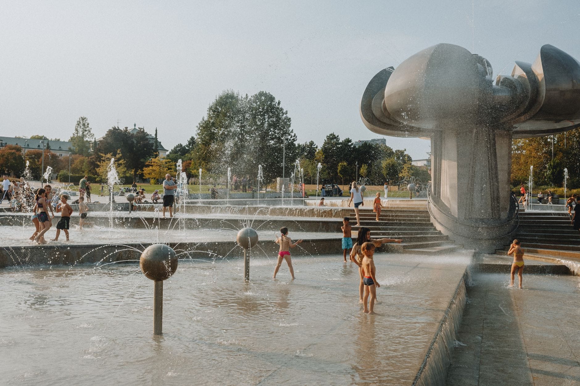 Children play in a large public fountain with modern sculpture in a green park setting in Prague on a sunny day.