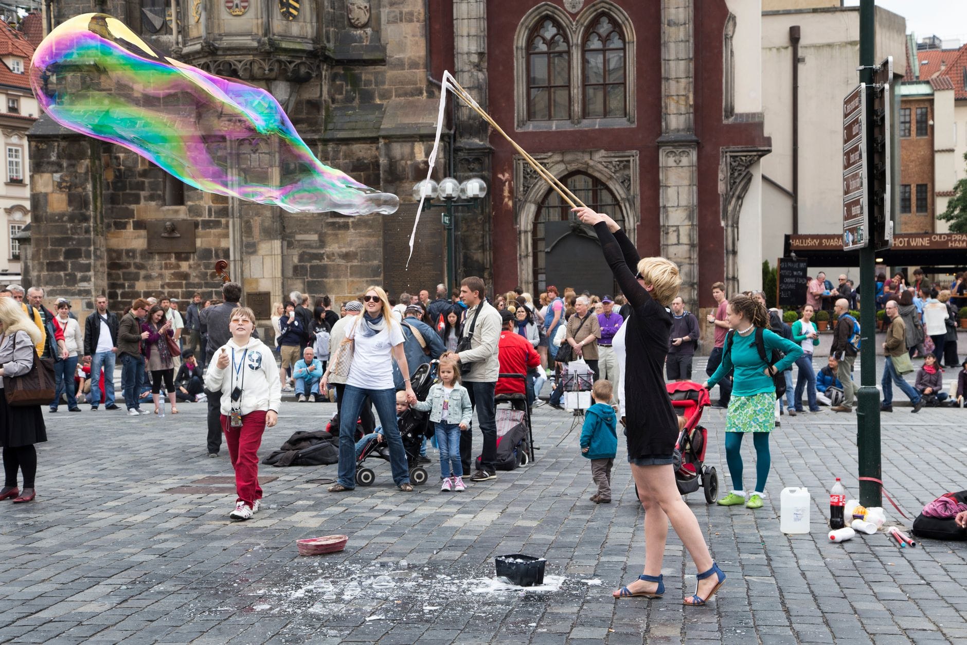 A lively city square in Prague with families, children, and a street performer making giant bubbles in front of historic buildings.