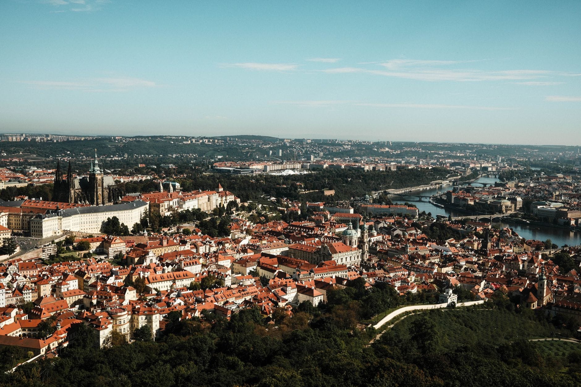 Wide panoramic view of Prague with red rooftops, historic buildings, and the Vltava river under a clear blue sky on a sunny day.