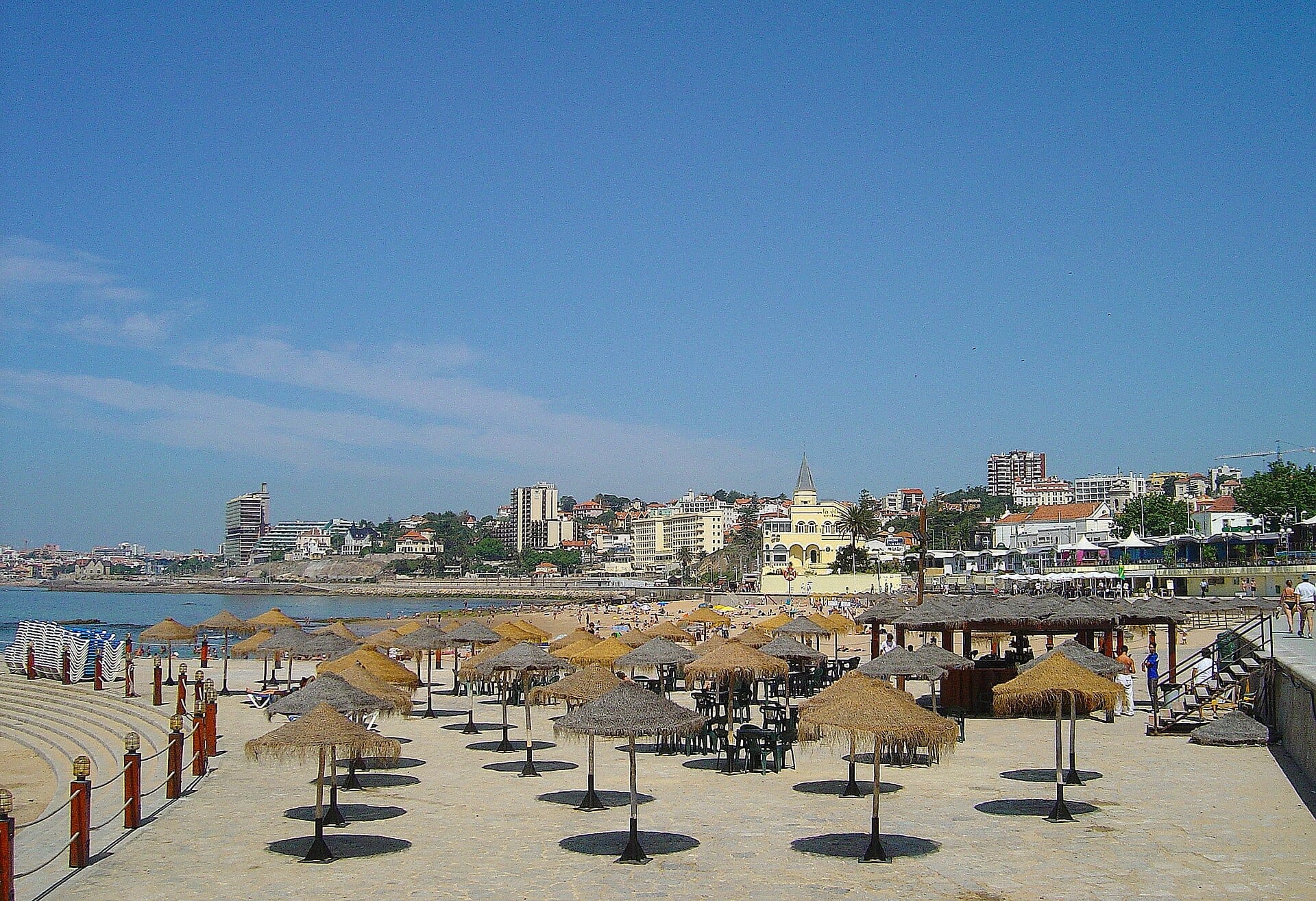 La spiaggia Praia do Tamariz a Estoril con file di ombrelloni di paglia, sabbia dorata ed edifici della città sotto un cielo azzurro limpido.
