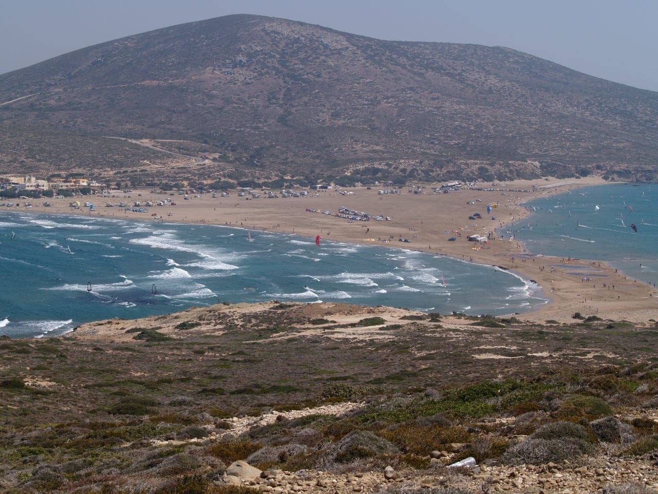 วิวกว้างของหาด Prasonisi ใน Rhodes ที่ซึ่งชายหาดทรายสองฝั่งและทะเลสีฟ้าบรรจบกัน พร้อมนักวินด์เซิร์ฟและนักท่องเที่ยวที่มองเห็นได้