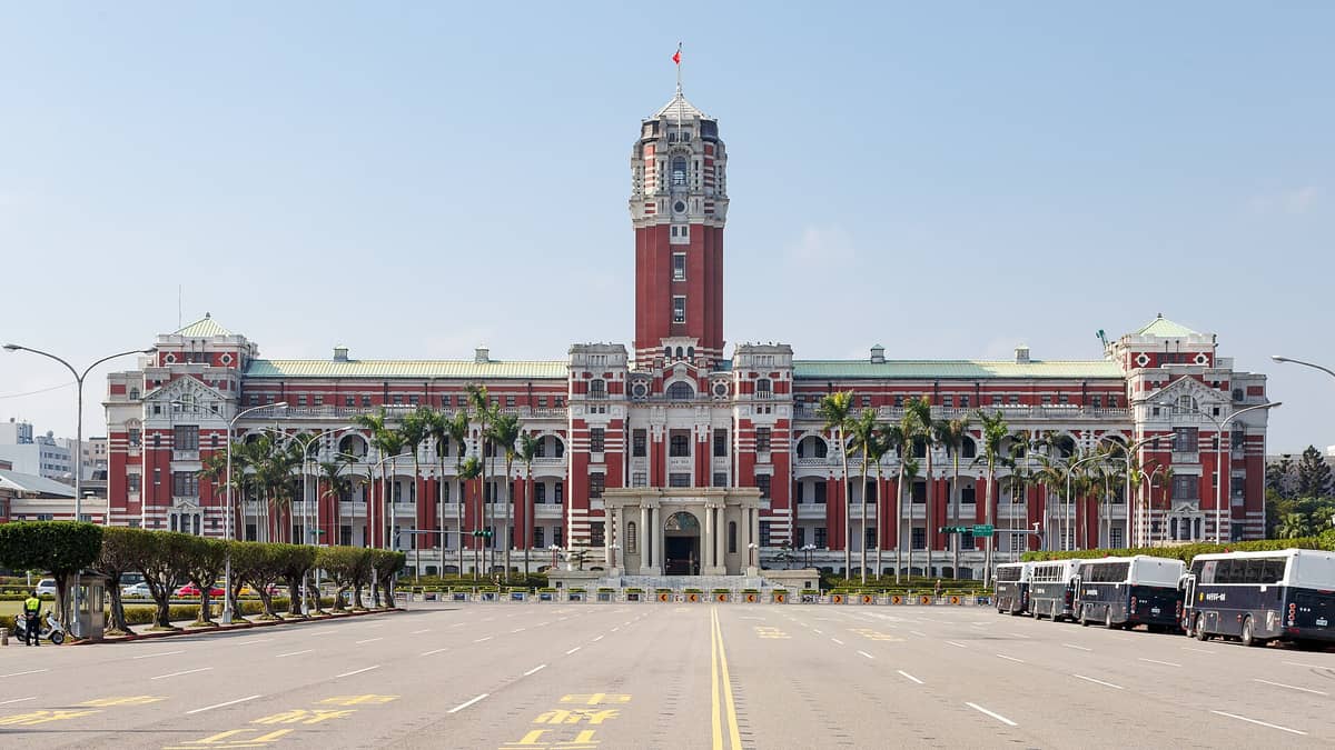 Sonnige Weitwinkelaufnahme des Taipeh Präsidialamts mit seiner Backsteinbarockfassade, dem zentralen Uhrenturm, Palmen und Bussen entlang des Boulevards.