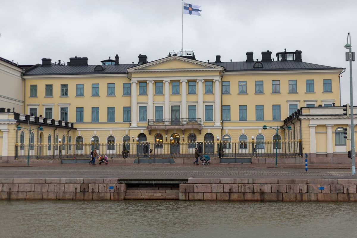 Vista frontal do Palácio Presidencial de Helsinque, um grandioso edifício neoclássico amarelo com grades de ferro e a bandeira finlandesa hasteada, visto a partir da orla marítima.