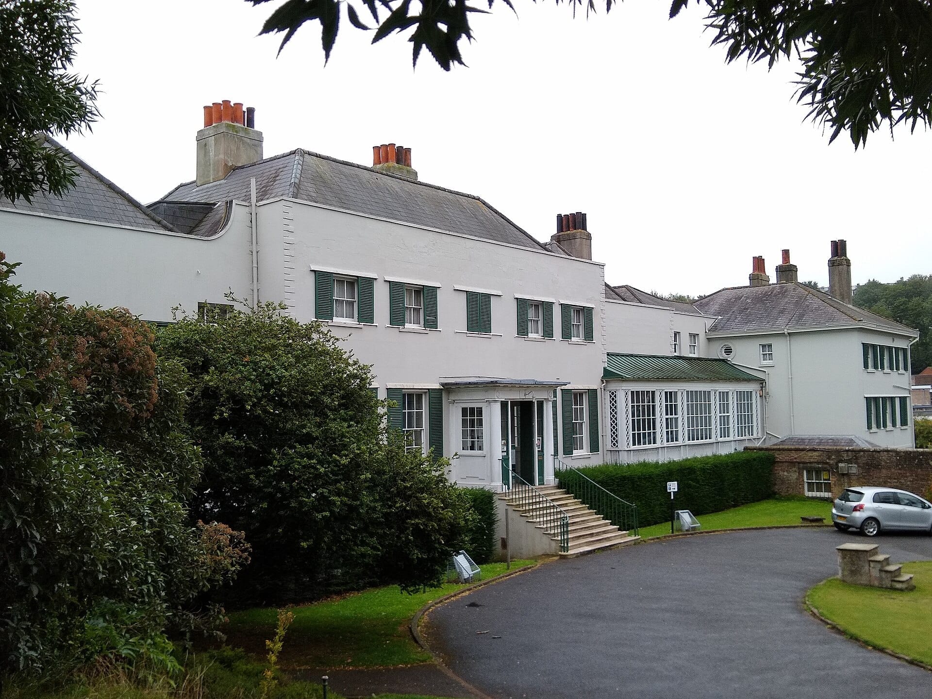 Preston Manor’s white Edwardian façade with green shutters, grand entrance steps, and garden greenery on a cloudy day in Brighton.