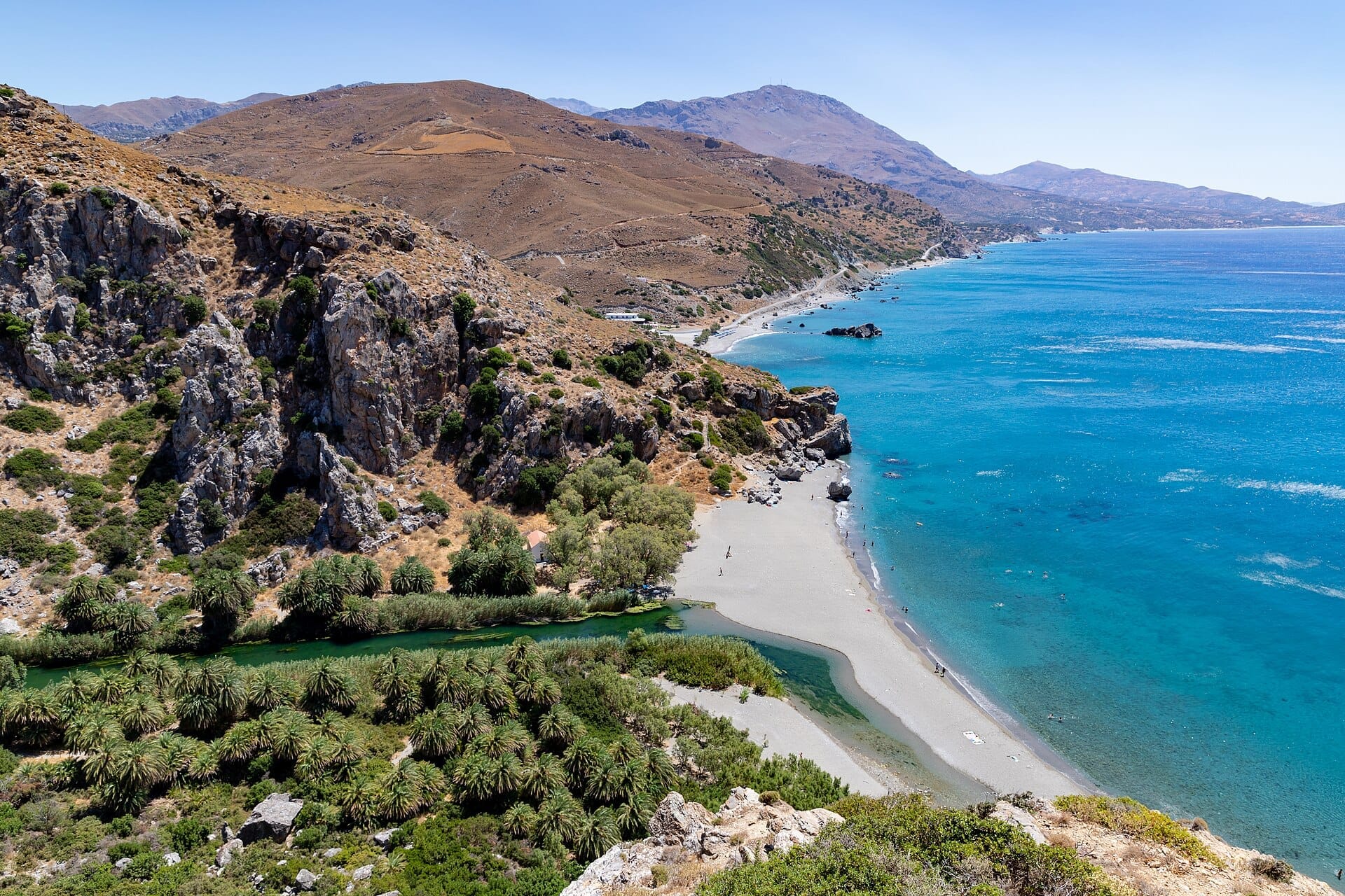 Vista panoramica della spiaggia di Preveli a Creta, con un boschetto di palme, acque turchesi, riva sabbiosa e montagne frastagliate.