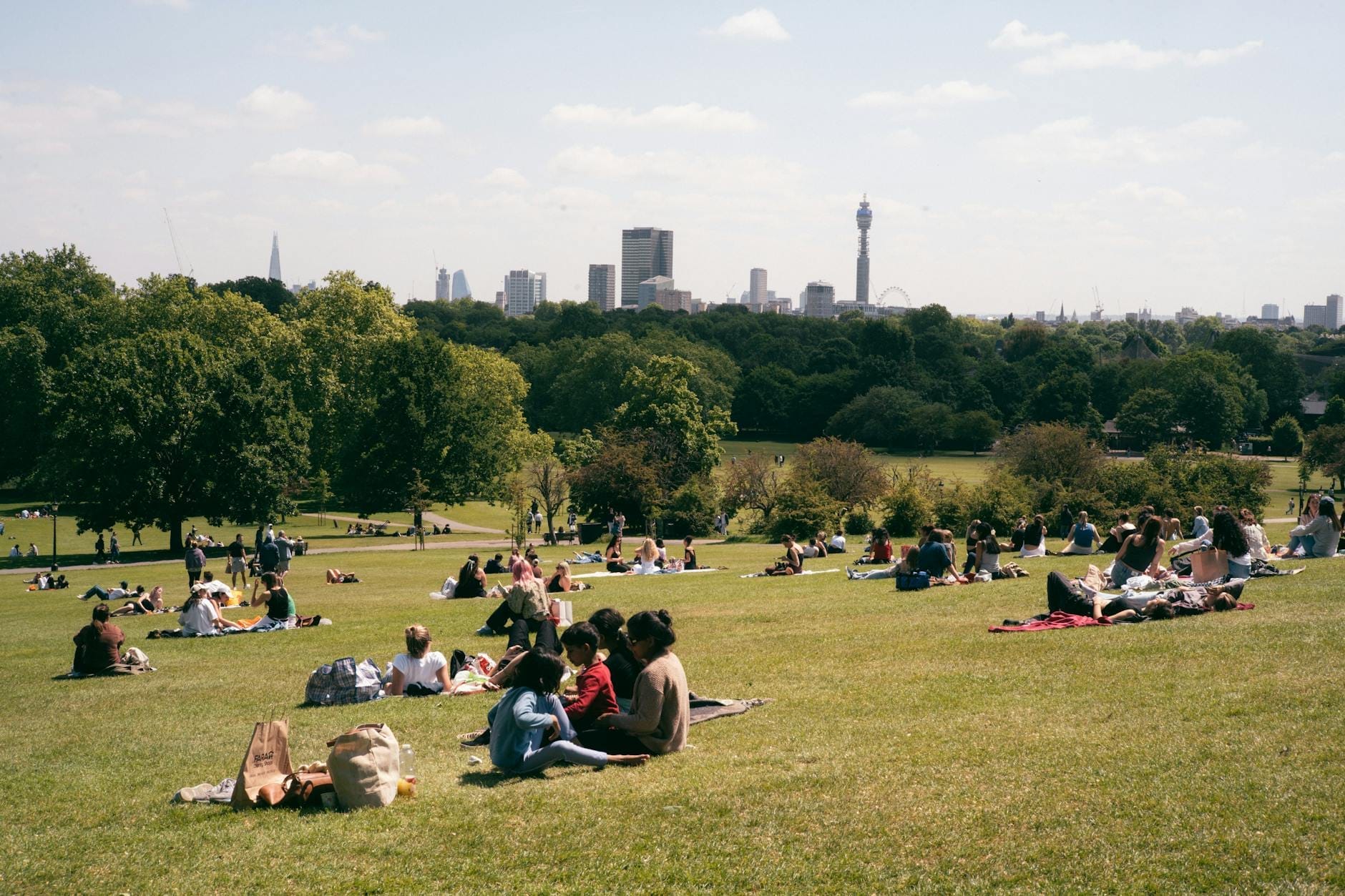 Persone distese sul prato di Primrose Hill con il panorama dello skyline di Londra e i suoi monumenti sullo sfondo, sotto un cielo terso.