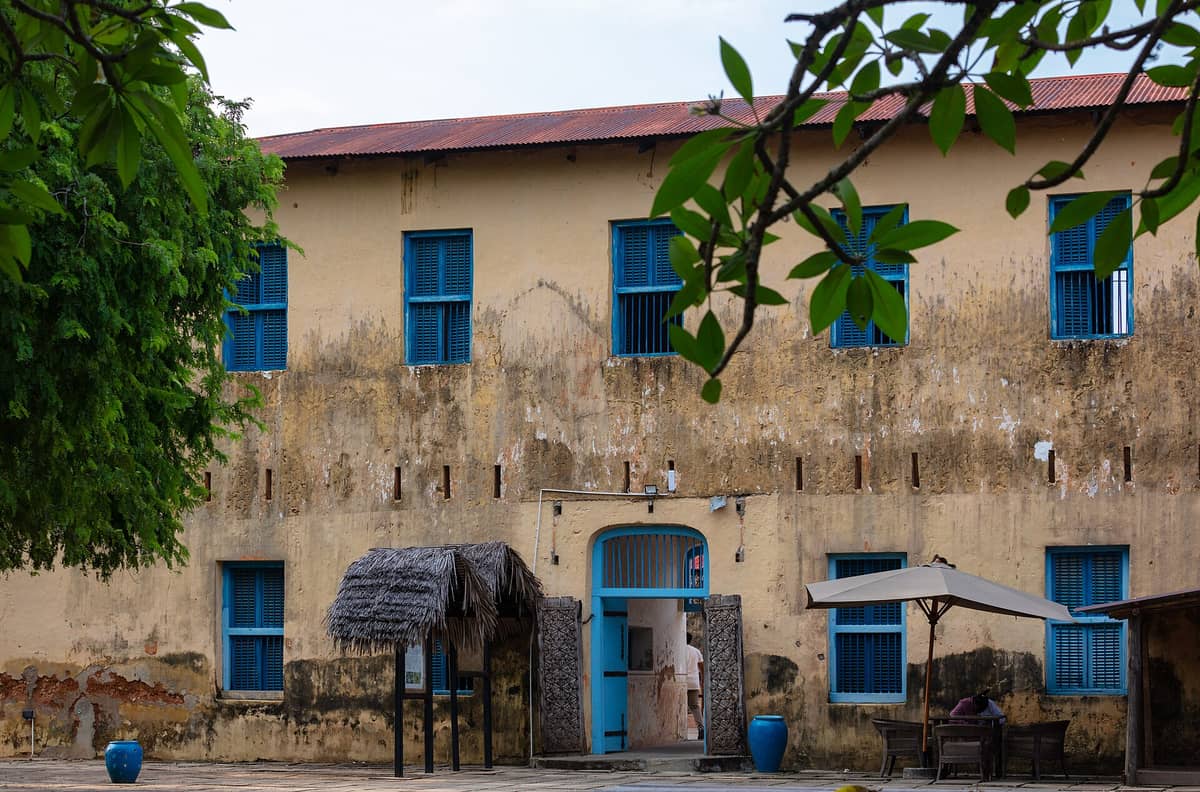 Historyczny żółty budynek więzienia z niebieskimi okiennicami na Prison Island (Wyspa Changuu) na Zanzibarze, częściowo otoczony drzewami i strzechą w pierwszym planie.