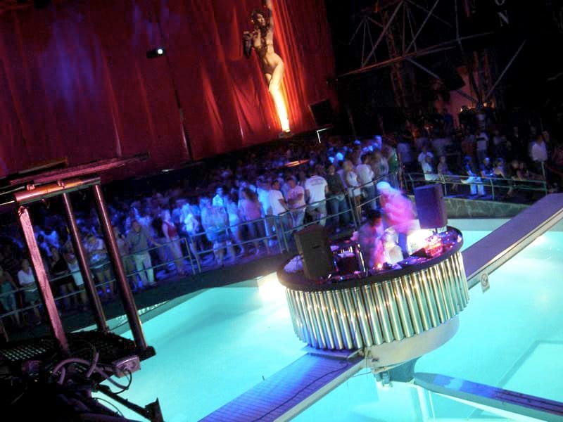 Grande folla all'interno della discoteca Privilege Ibiza, pista da ballo illuminata e cabina del DJ sopra una piscina, con drammatici drappeggi rossi sullo sfondo.